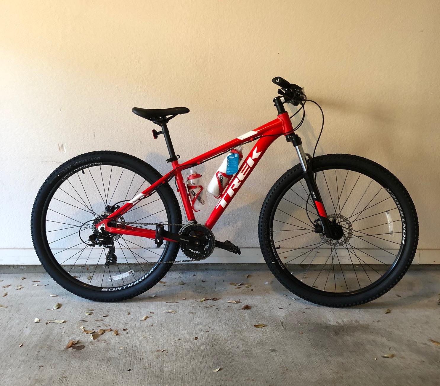 Trek Marlin 5 29er: A red Trek mountain bike is positioned against a neutral wall. The bike features thick tires, a front suspension fork, and a water bottle attached to the frame. Dry leaves are scattered on the ground beneath it.