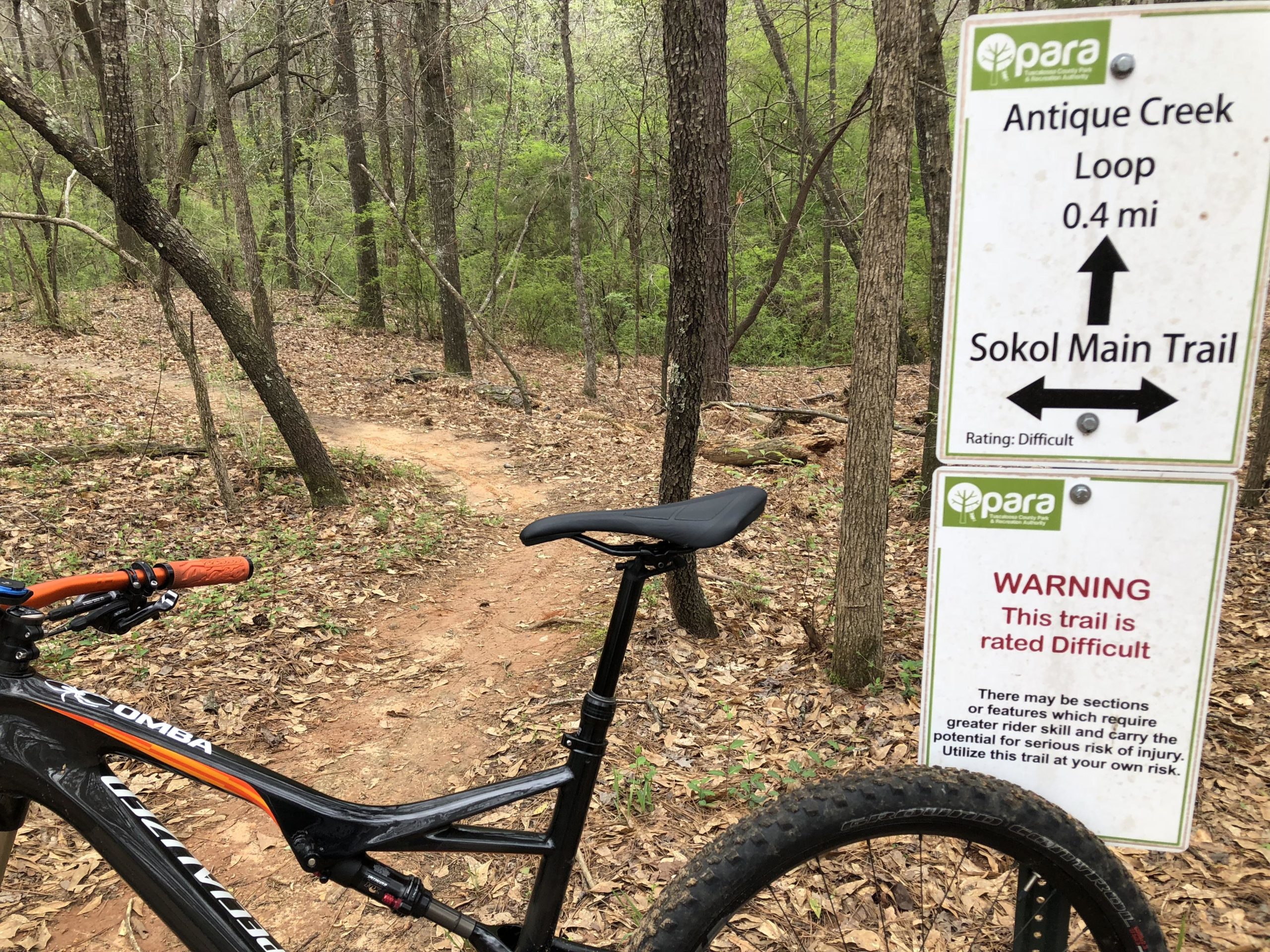 A black mountain bike is parked beside a trail sign in a wooded area. The sign indicates the Antique Creek Loop, measuring 0.4 miles, and the Sokol Main Trail, both rated as difficult. The surrounding landscape features bare trees and ground covered in leaves, suggesting a rustic and natural setting. Munny Sokol Park mountain bike trail.
