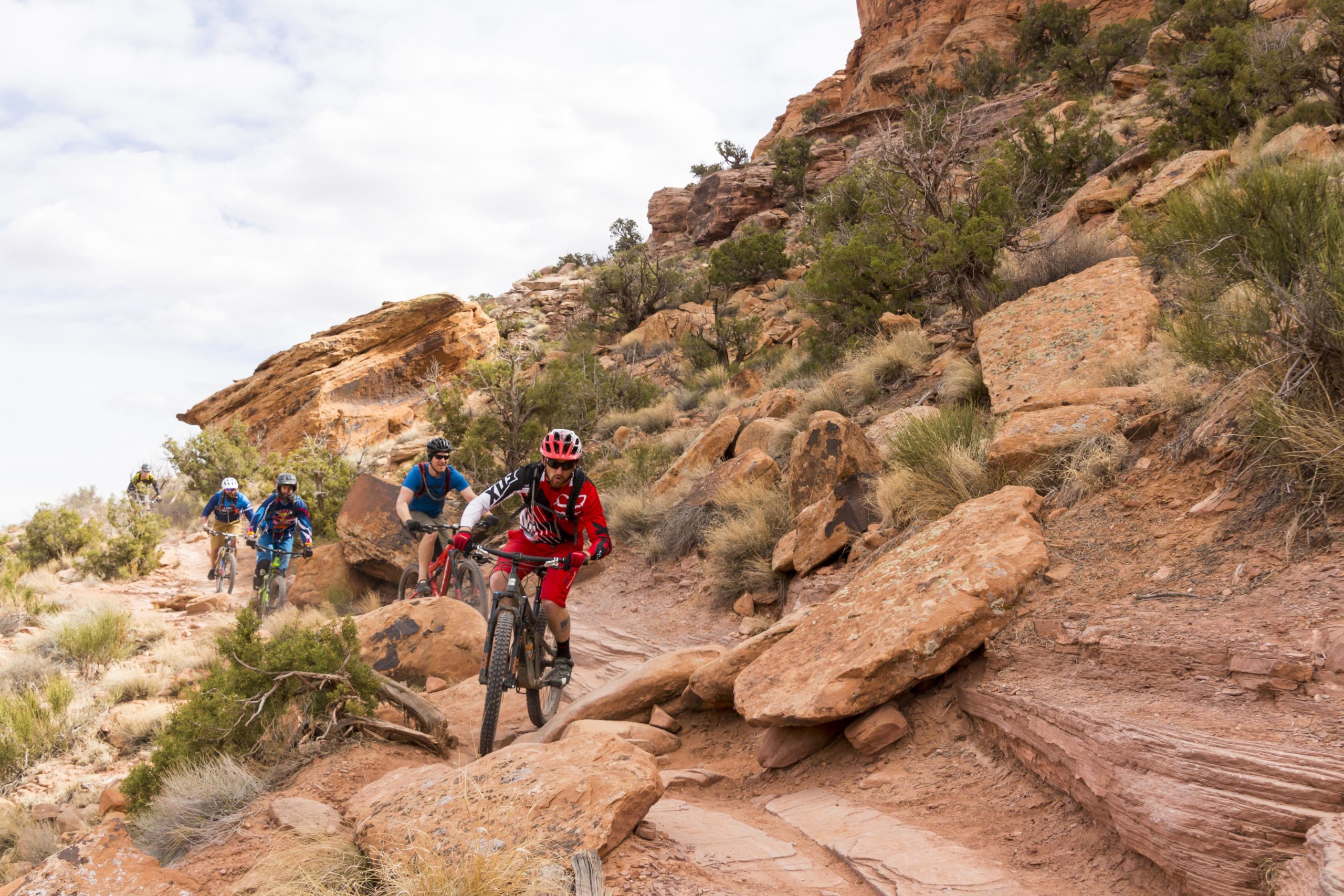 A group of four mountain bikers navigating a rocky trail in a desert landscape. The riders are wearing helmets and colorful biking gear, with two cyclists positioned in the foreground and two further back. The terrain features large rocks and sparse vegetation under a partly cloudy sky. Porcupine Rim mountain bike trail.