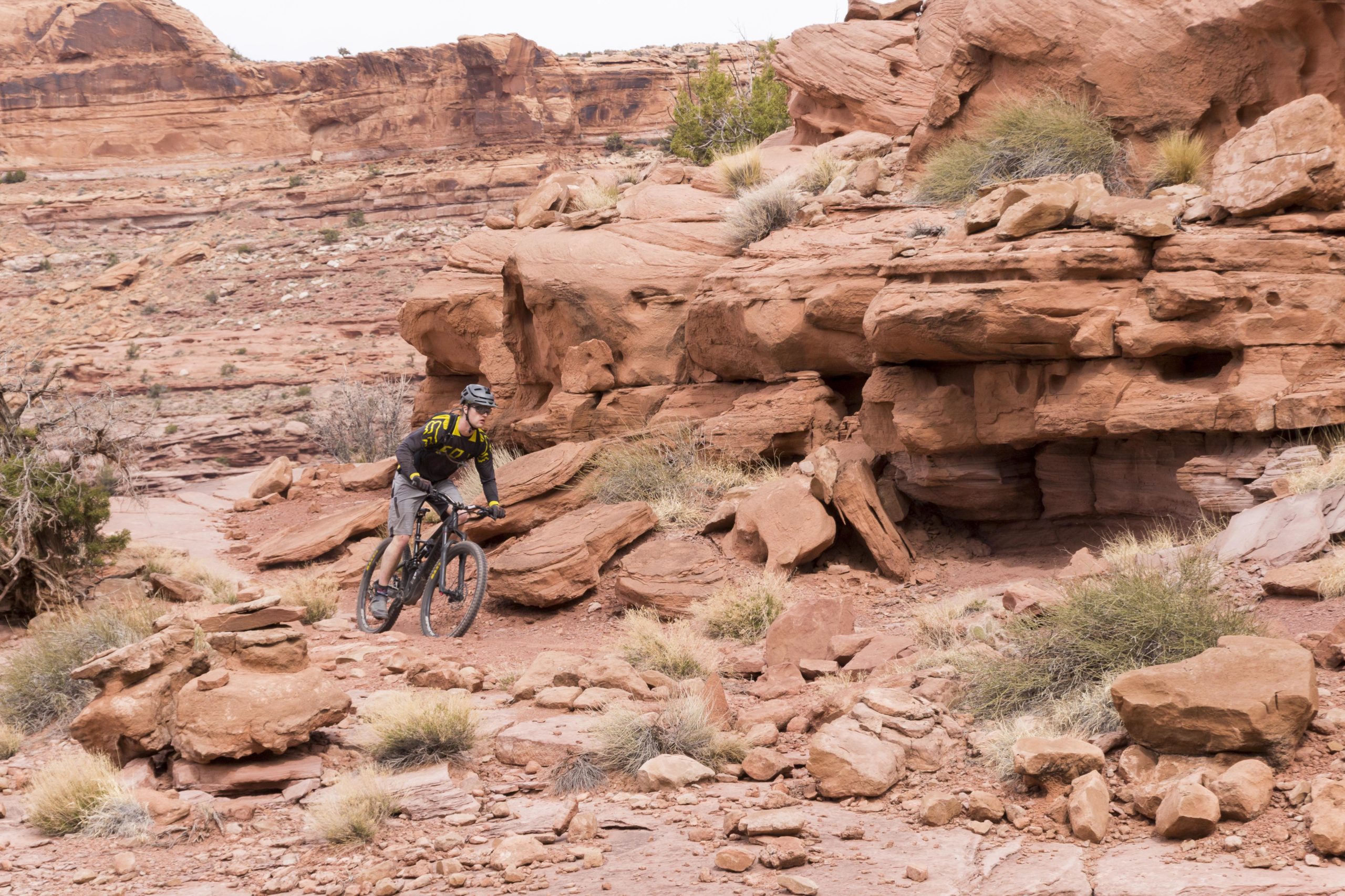 A mountain biker rides along a rocky trail in a desert landscape, surrounded by red rock formations and sparse vegetation. The cyclist is wearing protective gear, including a helmet and a brightly colored jersey, while navigating the uneven terrain. Porcupine Rim mountain bike trail.
