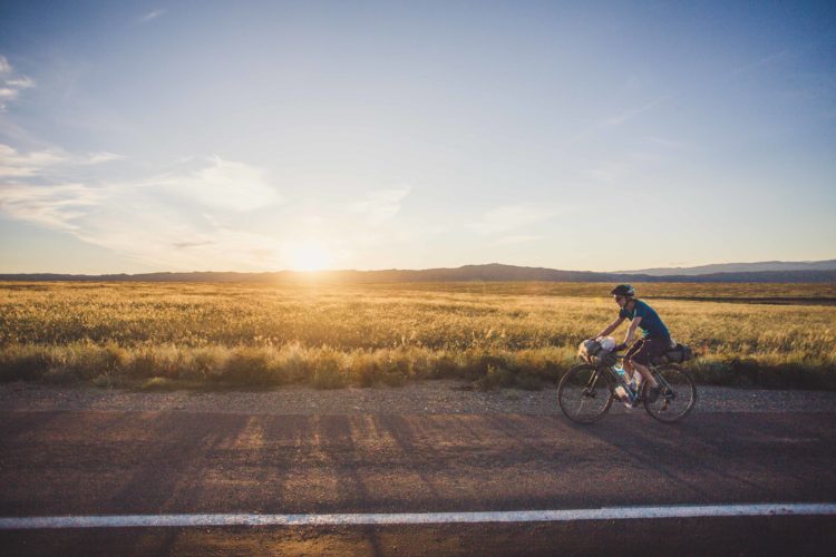 A cyclist riding along a rural road during sunset, with a vast field and mountains in the background. The sky is clear, displaying warm tones of orange and yellow as the sun sets on the horizon.