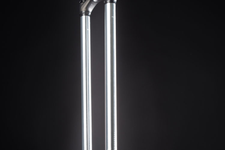A close-up view of a mountain bike front suspension fork, showcasing the shiny silver stanchions and black lower legs with mounting hardware against a black background.