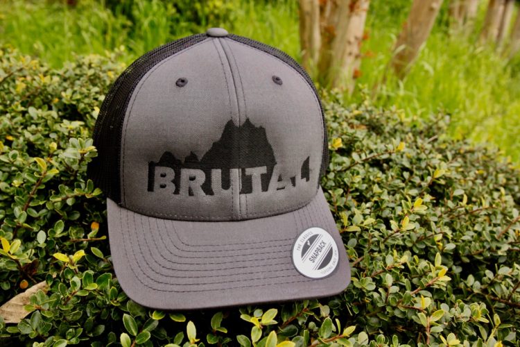 A gray snapback cap displayed on green foliage, featuring the word "BRUTA" in bold letters on the front alongside a mountain silhouette design. A sticker on the brim indicates it is part of "The Classics" collection.