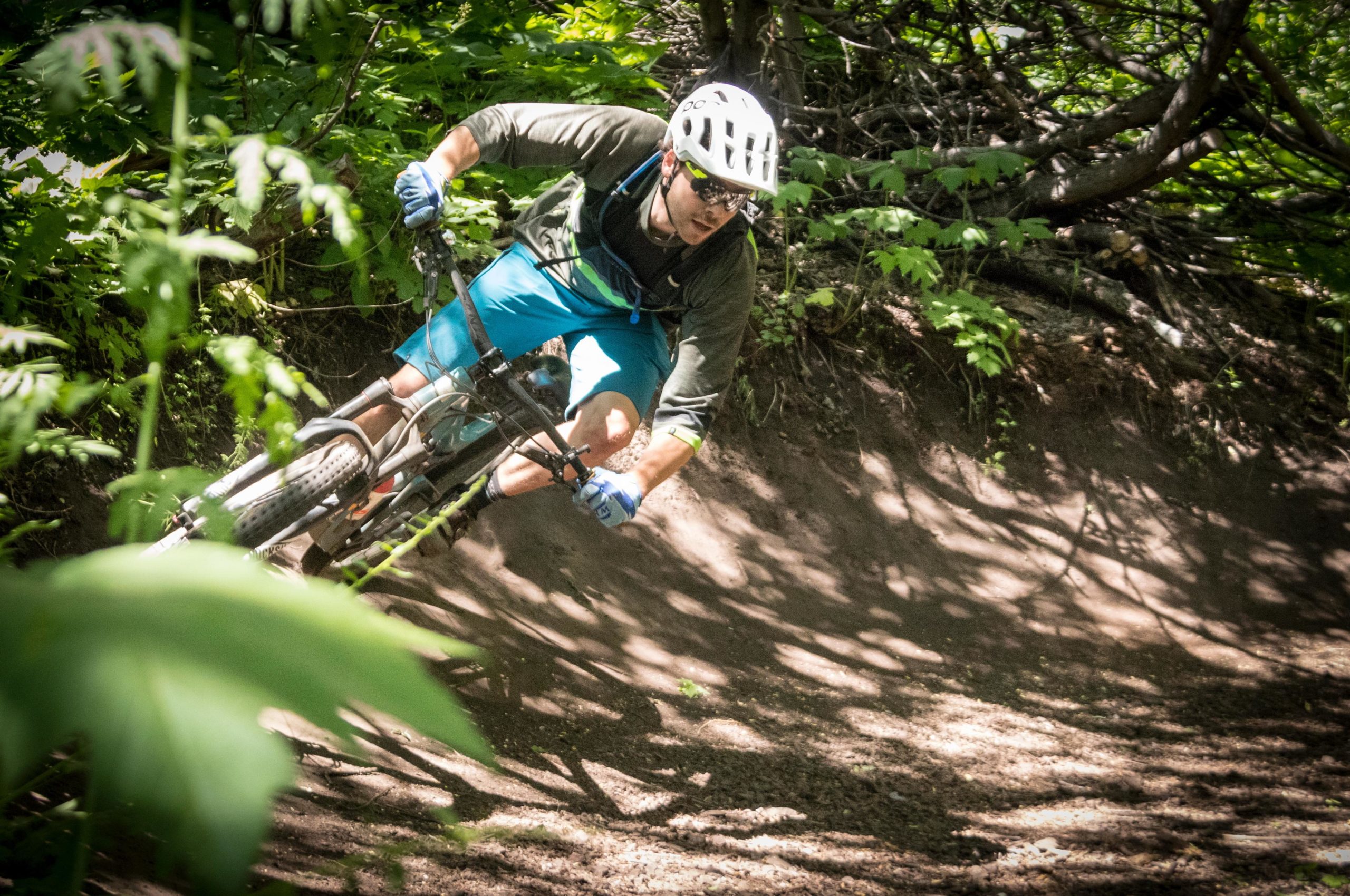A mountain biker navigating a dirt path in a lush green forest, leaning into a turn while wearing a helmet, sunglasses, and biking gloves. Sunlight filters through the trees, creating dappled shadows on the ground. W.O.W Trail mountain bike trail.