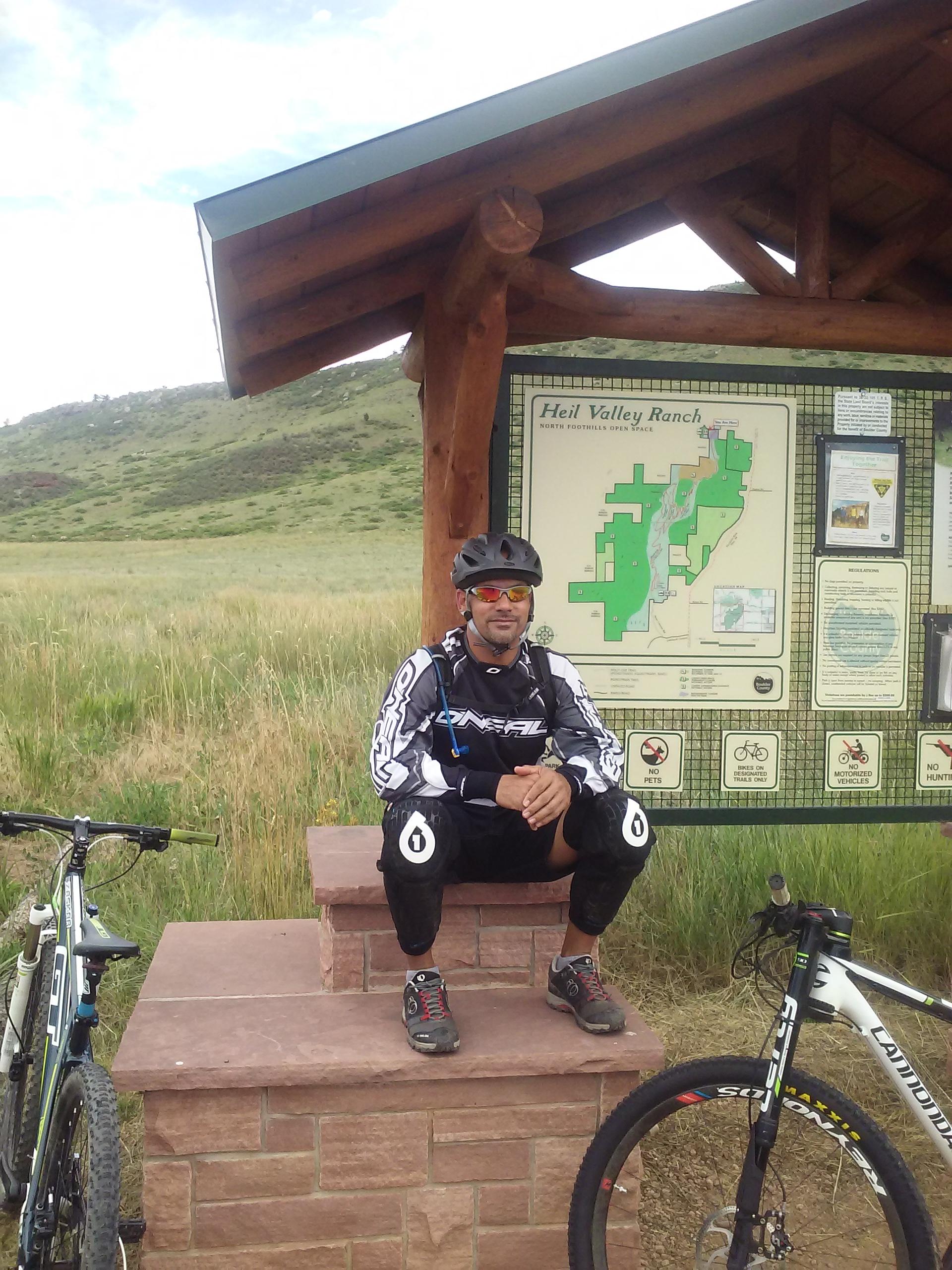 A person wearing a helmet and biking gear sits on a stone platform near a trailhead sign for Heil Valley Ranch. Two mountain bikes are parked beside them, with green hills and grass in the background under a partly cloudy sky. Heil Valley Ranch mountain bike trail.
