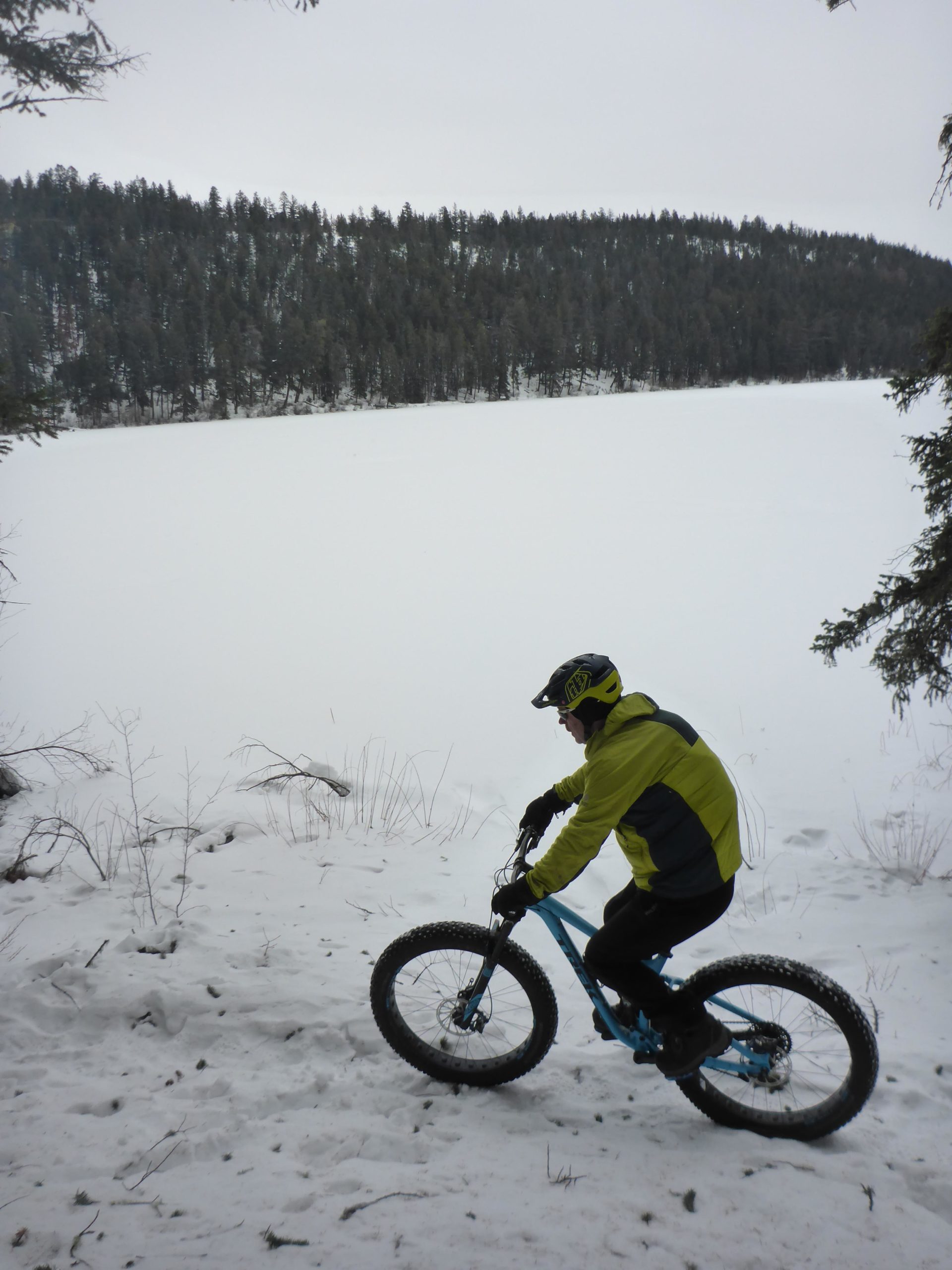 A person riding a fat bike along a snow-covered path near a frozen lake, surrounded by evergreen trees. The cyclist is wearing a bright yellow and gray jacket, gloves, and a helmet, with overcast skies in the background. Pyramid Bench mountain bike trail.