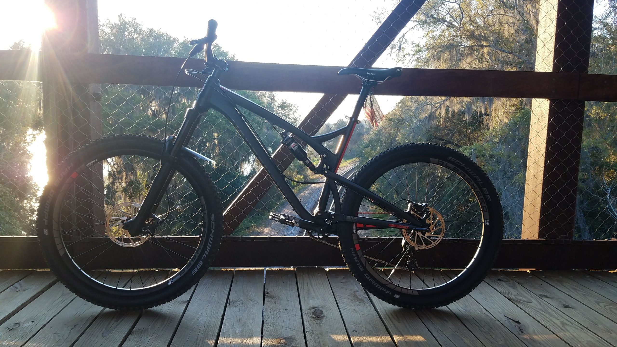 A mountain bike parked on a wooden bridge with a scenic view of trees and water in the background, illuminated by the sun setting in the distance. The bike features wide tires and a modern frame design. Tom Brown / Lafayette Heritage Park mountain bike trail.