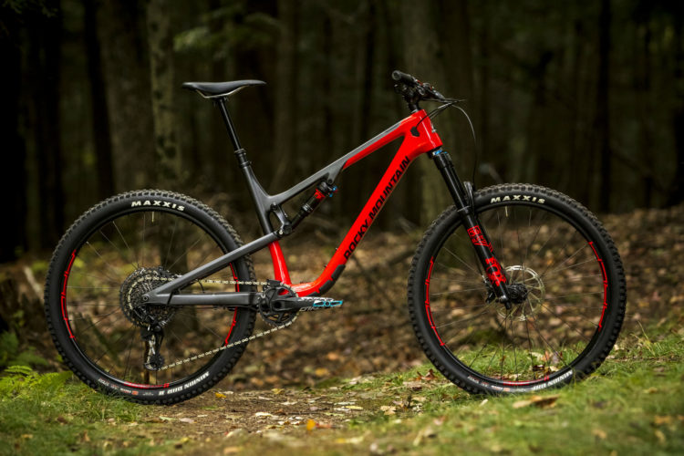 A modern mountain bike with a grey and red frame, featuring Maxxis tires, positioned on a wooded trail with greenery and fallen leaves in the background.