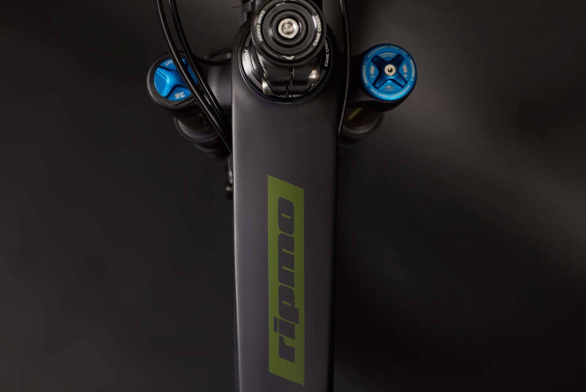 Ibis RipMo: Close-up view of a bicycle frame featuring the word "Ripmo" in green. The image shows handlebars with blue adjustment knobs and parts of the front suspension. The background is dark, emphasizing the bike's sleek design.