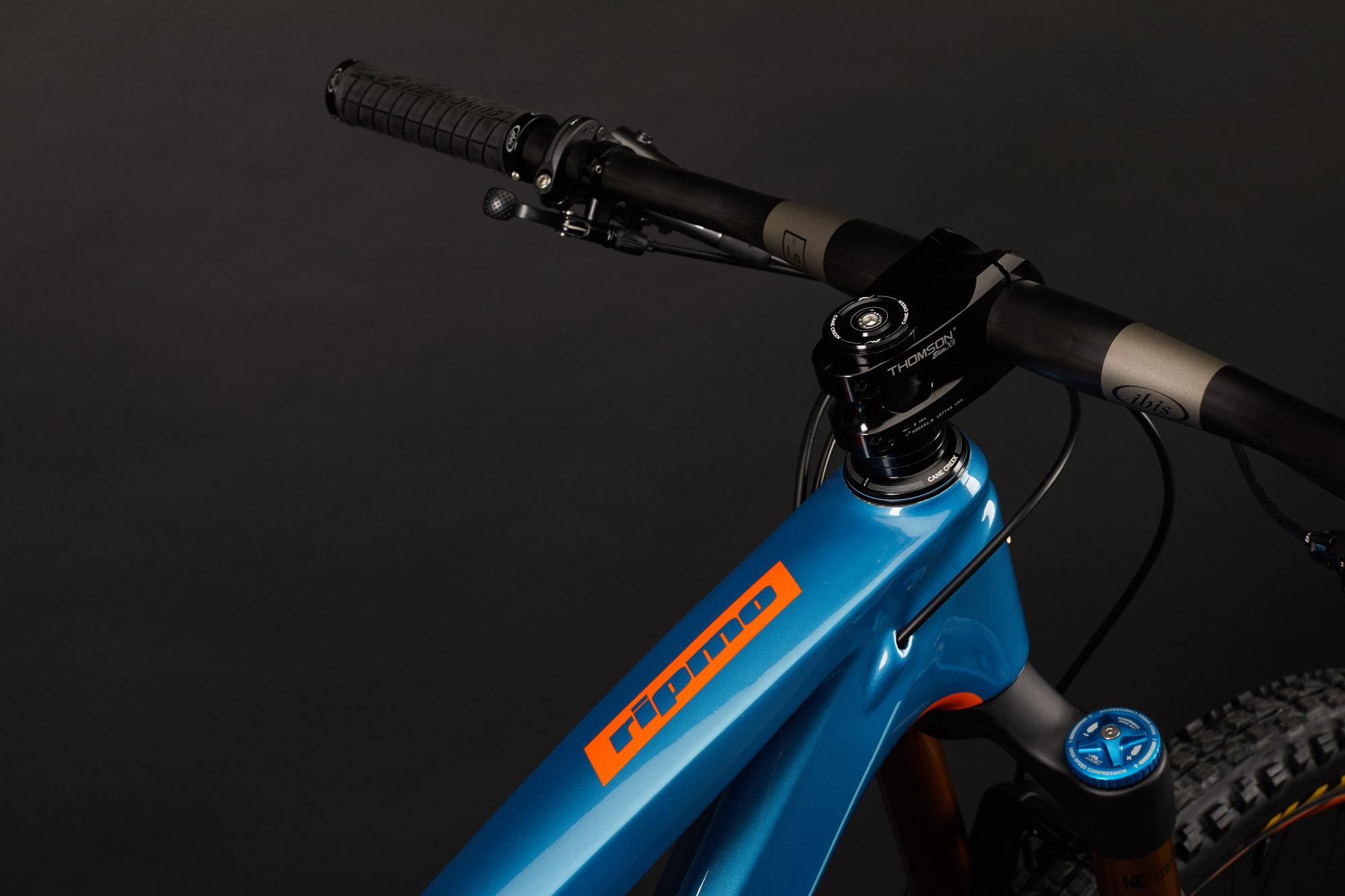 Ibis RipMo: Close-up view of the handlebars and top tube of a blue mountain bike, featuring a prominent orange logo. The bike's stem, fork, and detailed grip can be seen against a dark background, highlighting its sleek design and components.