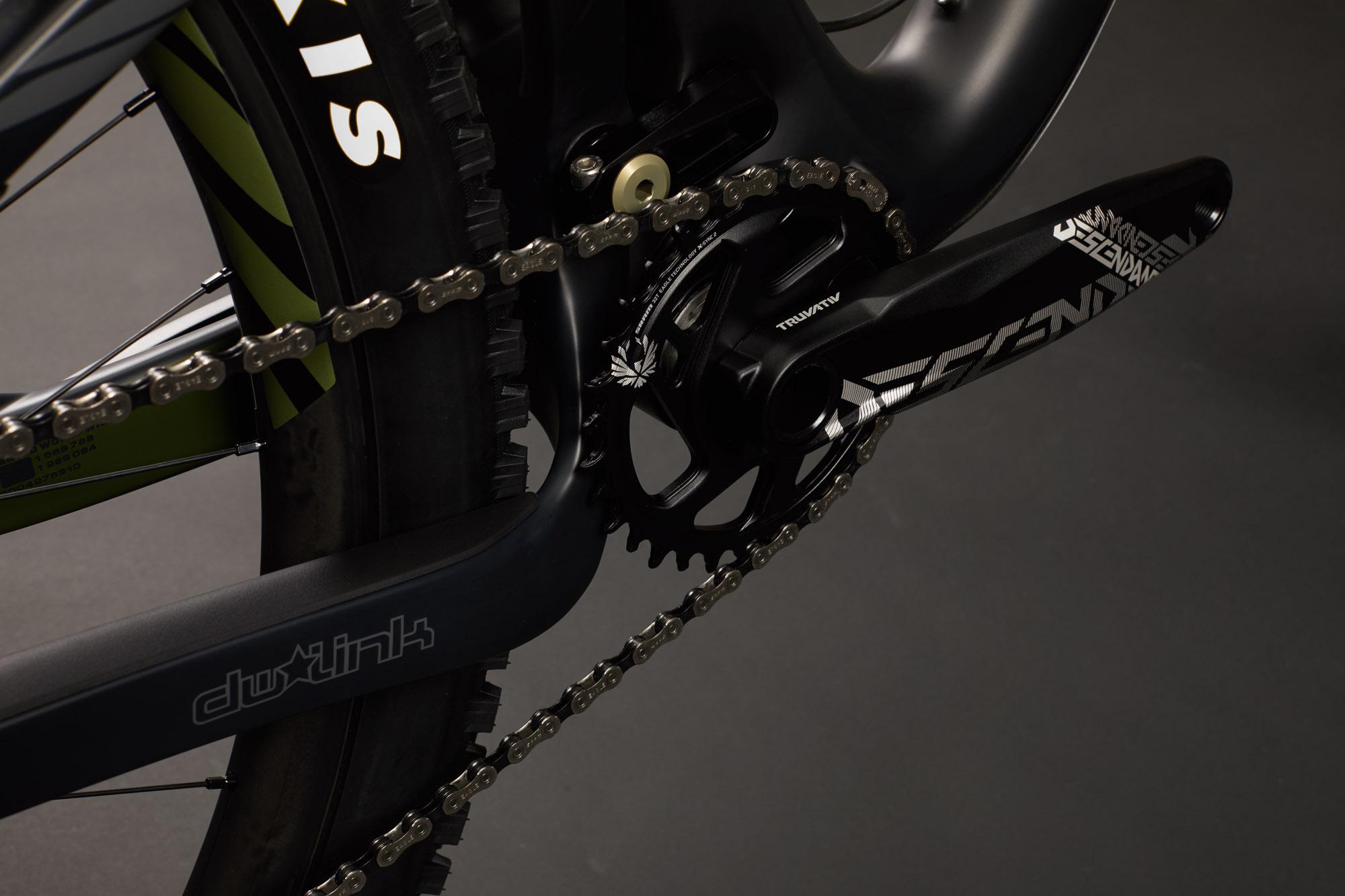 Ibis RipMo: Close-up view of a bicycle's drivetrain, showcasing the chain, crankset, and pedal. The components are detailed against a dark background, highlighting the intricate design and branding of the parts, including a prominent chainring and crank arm.