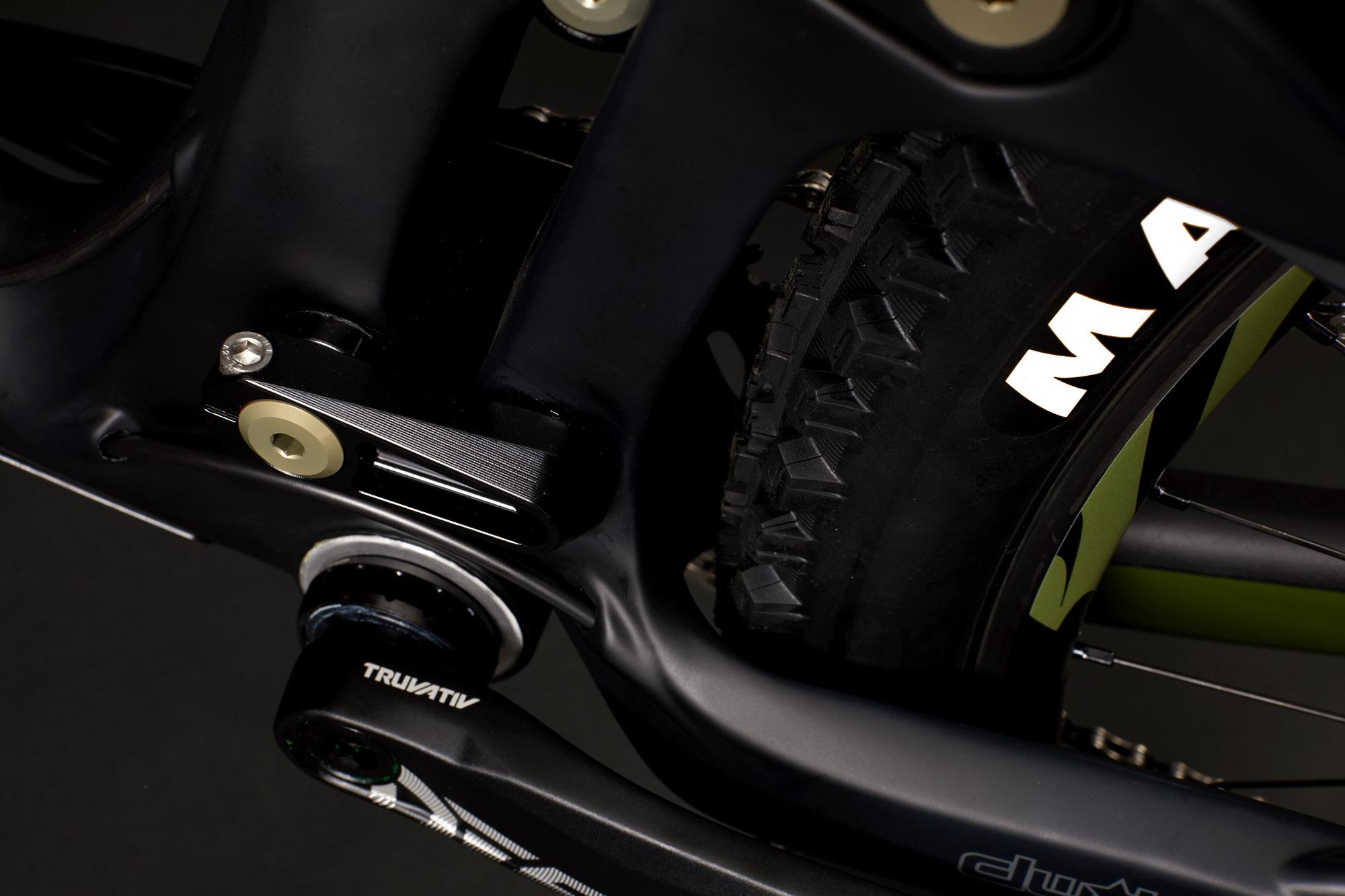 Ibis RipMo: Close-up of a mountain bike's rear suspension system, showcasing the drivetrain and tire. The image highlights a black frame component labeled "TRUVATIV," the glossy rear tire with visible tread, and a smooth metal part with a gold accent. The background is dark, emphasizing the bike's features.