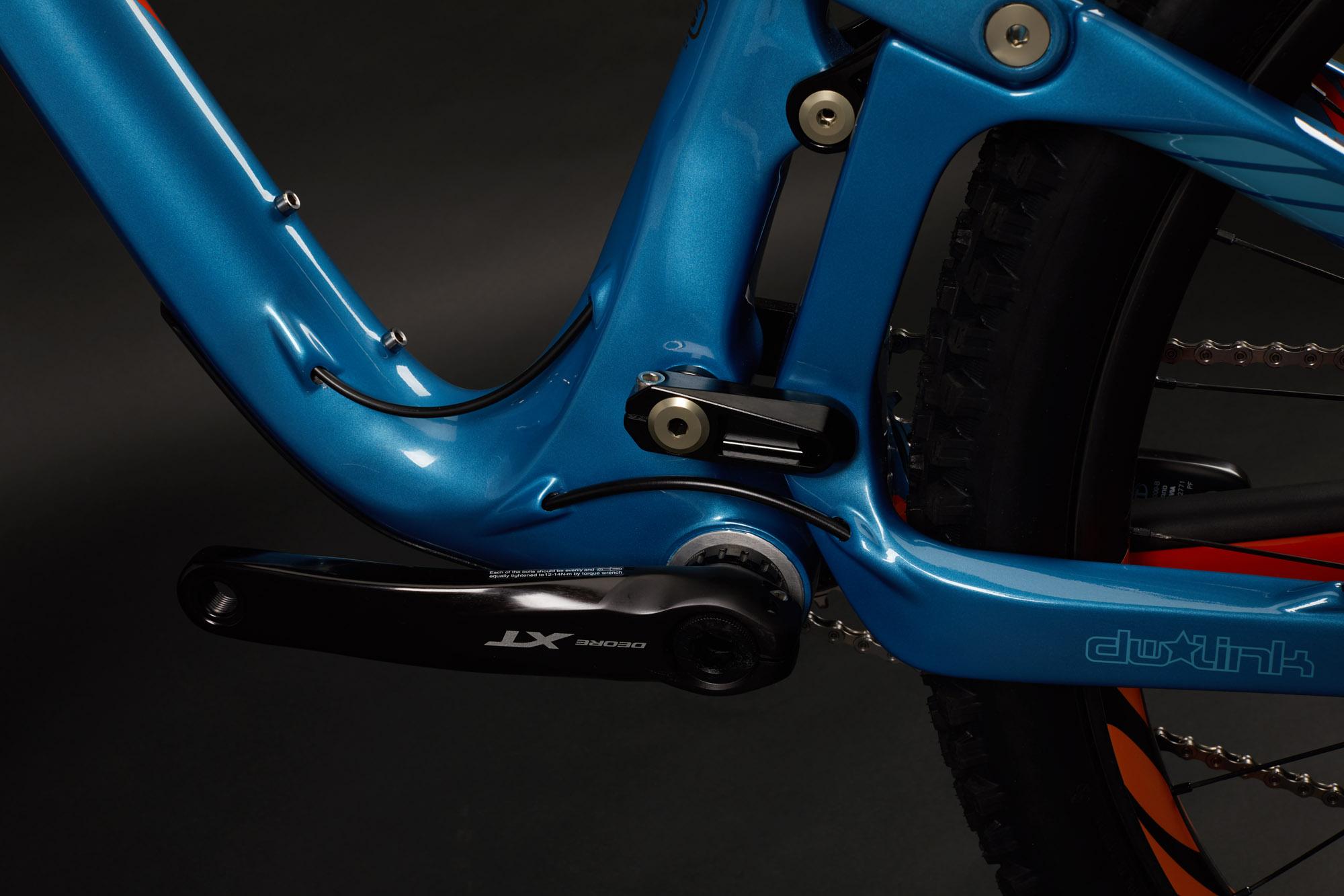 Ibis RipMo: Close-up view of a blue bicycle frame with a focus on the bottom bracket area and cranks. The frame features a shiny finish and is equipped with Shimano XT components. The background is dark, highlighting the bike's details, including the intricate linkage design.