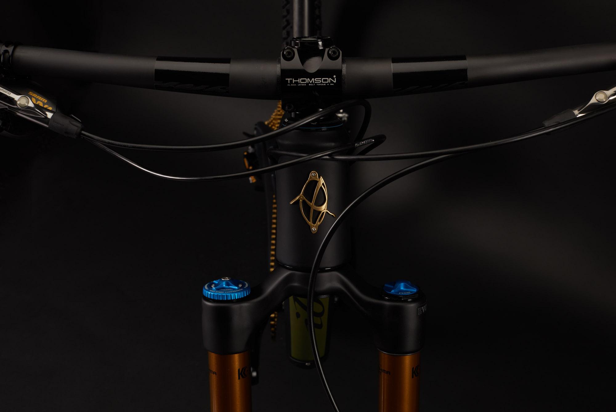 Ibis RipMo: Close-up view of a mountain bike's front end, featuring a black handlebar with a Thomson stem, cables neatly routed, and a distinctive gold logo on the frame. The bike's suspension forks have blue adjustment knobs, and the background is a solid black for emphasis.