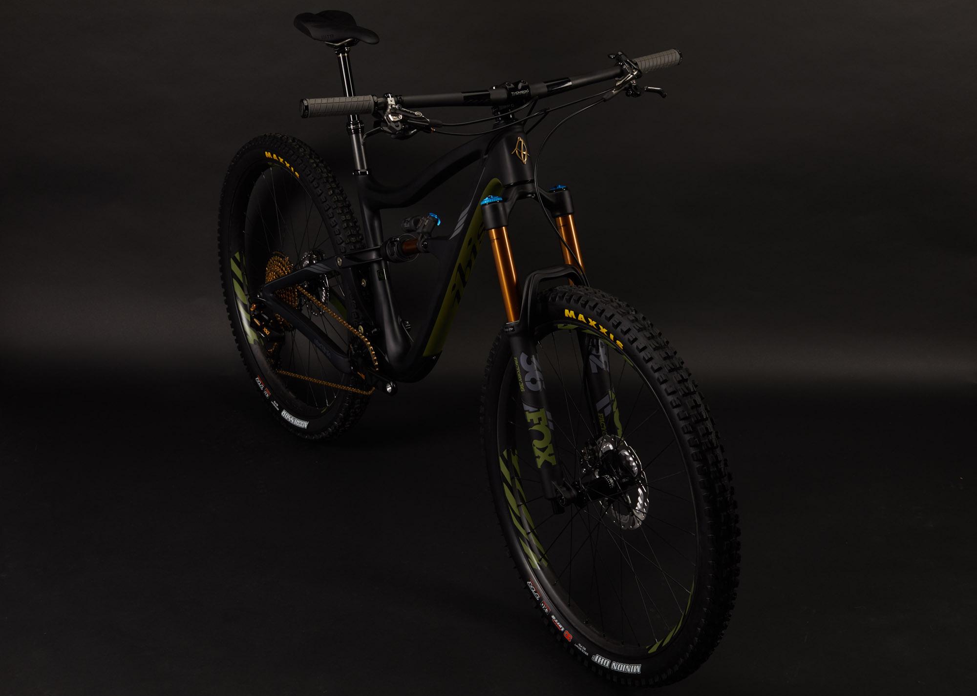 Ibis RipMo: A modern mountain bike with a sleek, matte black and green frame, featuring prominent front suspension with orange accents. The bike has wide, knobby tires suitable for off-road terrain, and the handlebars are equipped with textured grips. The background is dark, emphasizing the bike's design and detailing.