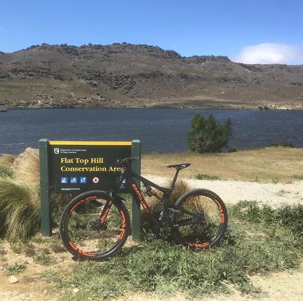 Santa Cruz Bronson C: A black mountain bike with orange accents leaning against a green sign that reads "Flat Top Hill Conservation Area." Behind the sign, there is a body of water and rolling hills under a clear blue sky.