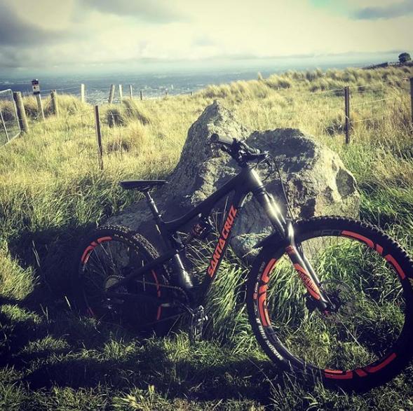 Santa Cruz Bronson C: A black mountain bike with orange accents rests against a large rock in a grassy field, overlooking a scenic landscape under a partly cloudy sky.