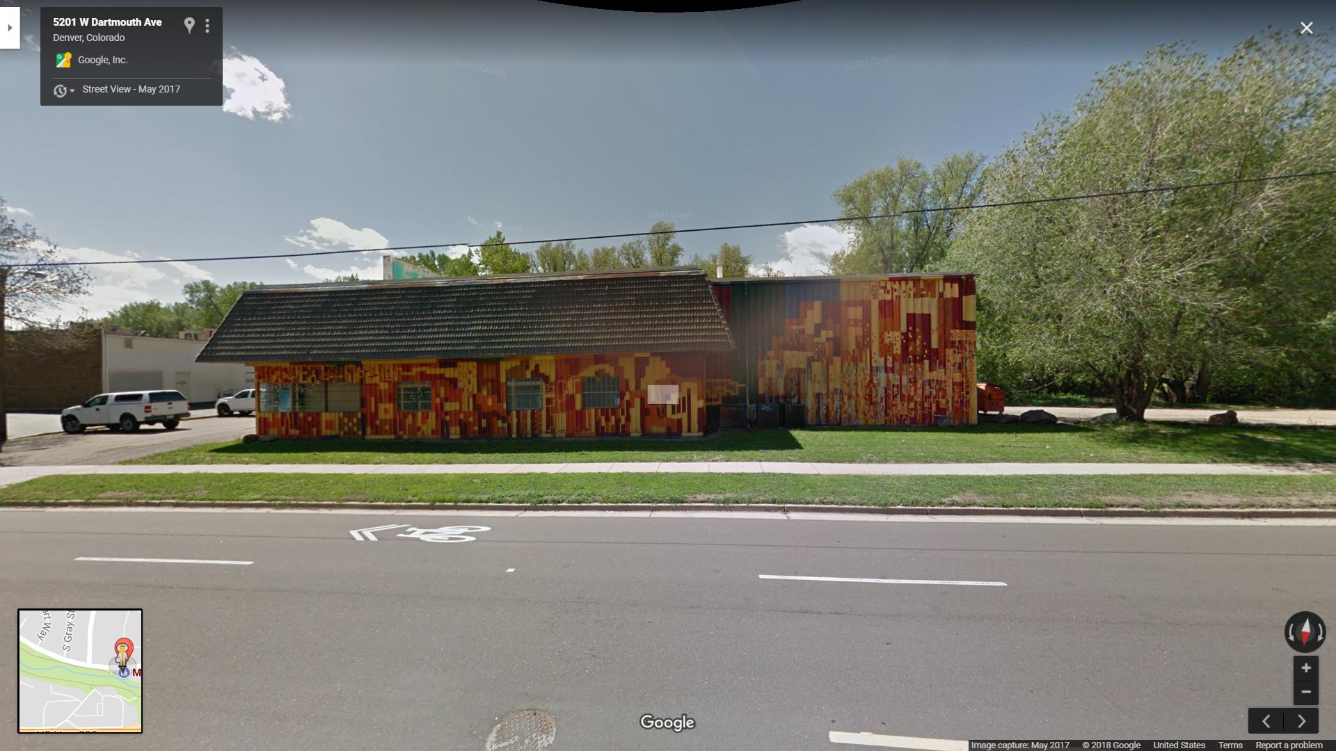 A colorful, mural-covered building located at 5201 W Dartmouth Ave in Denver, Colorado, viewed from across the street. The structure features a combination of orange, red, and yellow patterns. A grassy area and tree are visible in the foreground, along with a bicycle lane along the road.