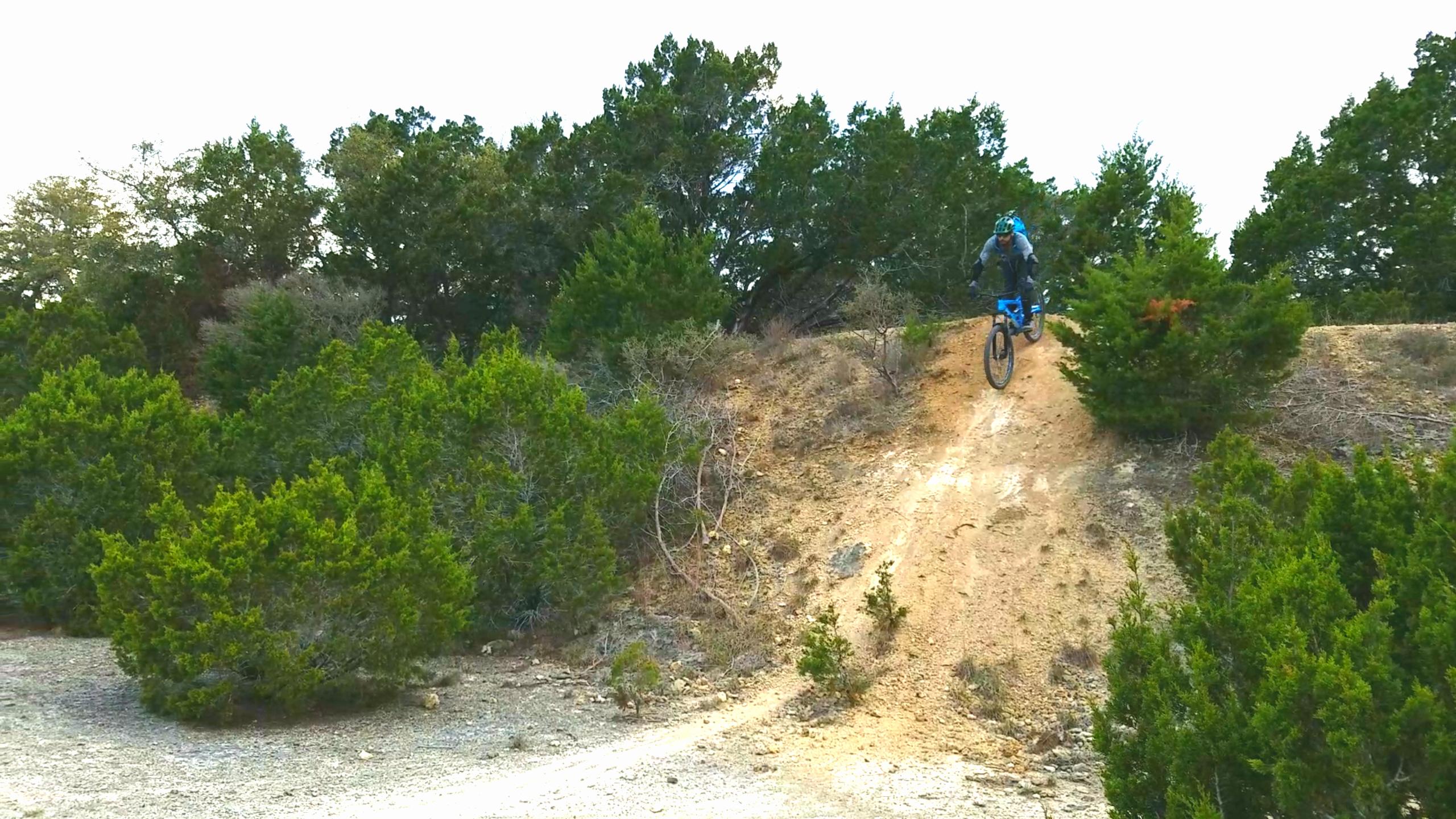 A mountain biker performing a jump on a dirt trail surrounded by greenery and trees. The rider is dressed in blue and gray, soaring off a mound of dirt with a scenic background of bushes and trees. Pace Bend Park mountain bike trail.