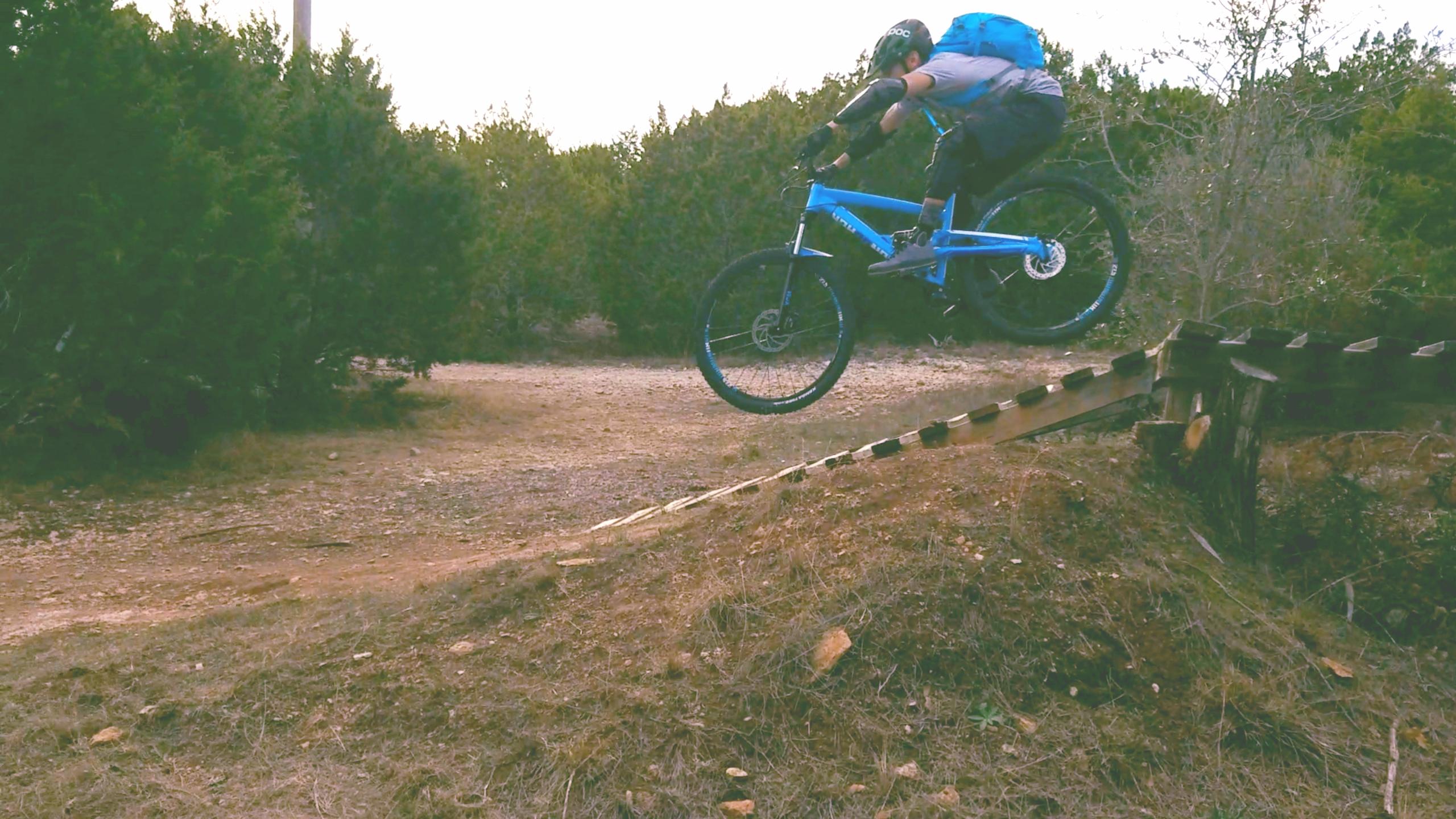 A mountain biker in protective gear jumps off a wooden ramp, soaring through the air above a dirt pathway surrounded by trees. The bike is blue, and the rider is wearing a helmet and a backpack, showcasing a dynamic, action-packed moment in a natural outdoor setting. Pace Bend Park mountain bike trail.