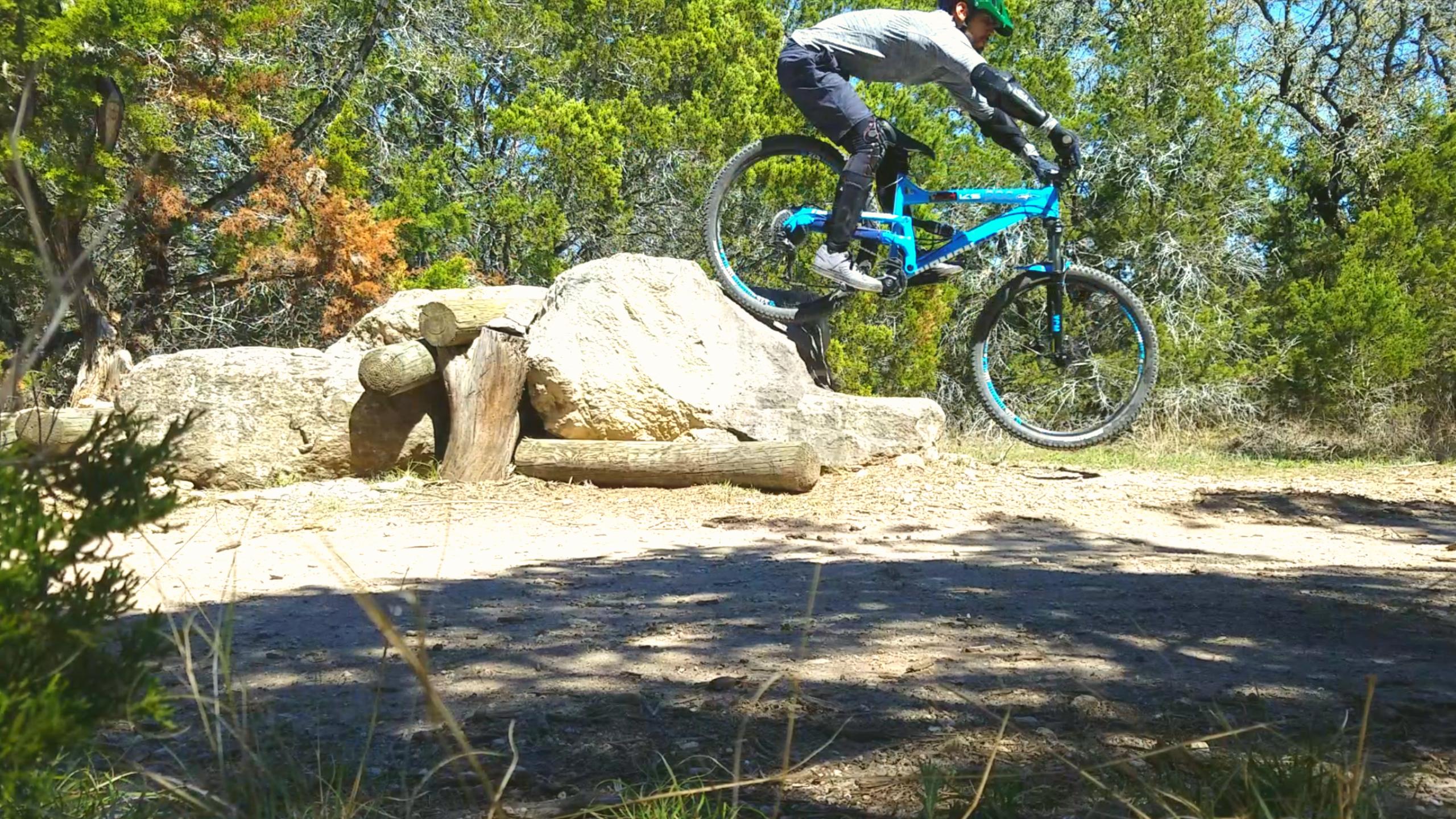 A mountain biker jumps off a rock formation on a dirt trail surrounded by trees, showcasing an energetic stunt with vibrant blue mountain bike gear. Pace Bend Park mountain bike trail.
