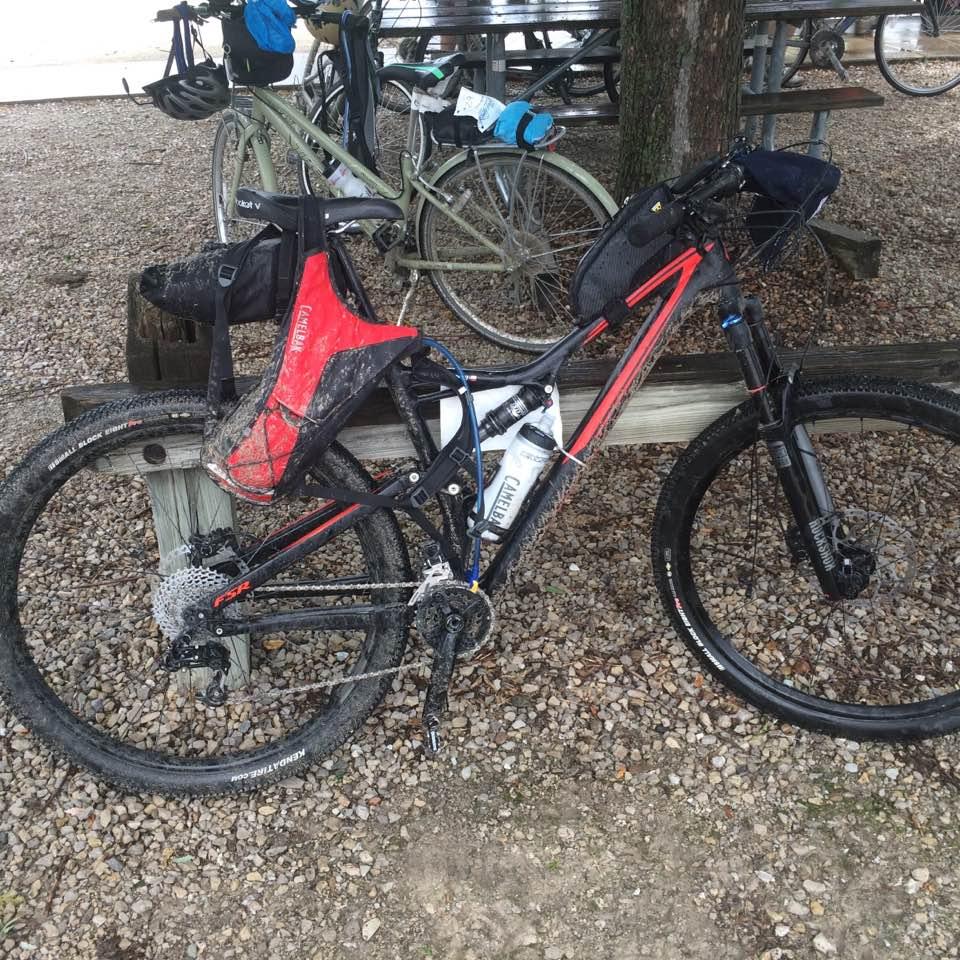 Specialized Stumpjumper: A mountain bike with a red saddle bag and a water bottle attached, parked beside a wooden beam on a gravel surface. Other bicycles are visible in the background, alongside a picnic area covered by trees. The bike shows signs of mud, indicating recent use on rugged terrain.