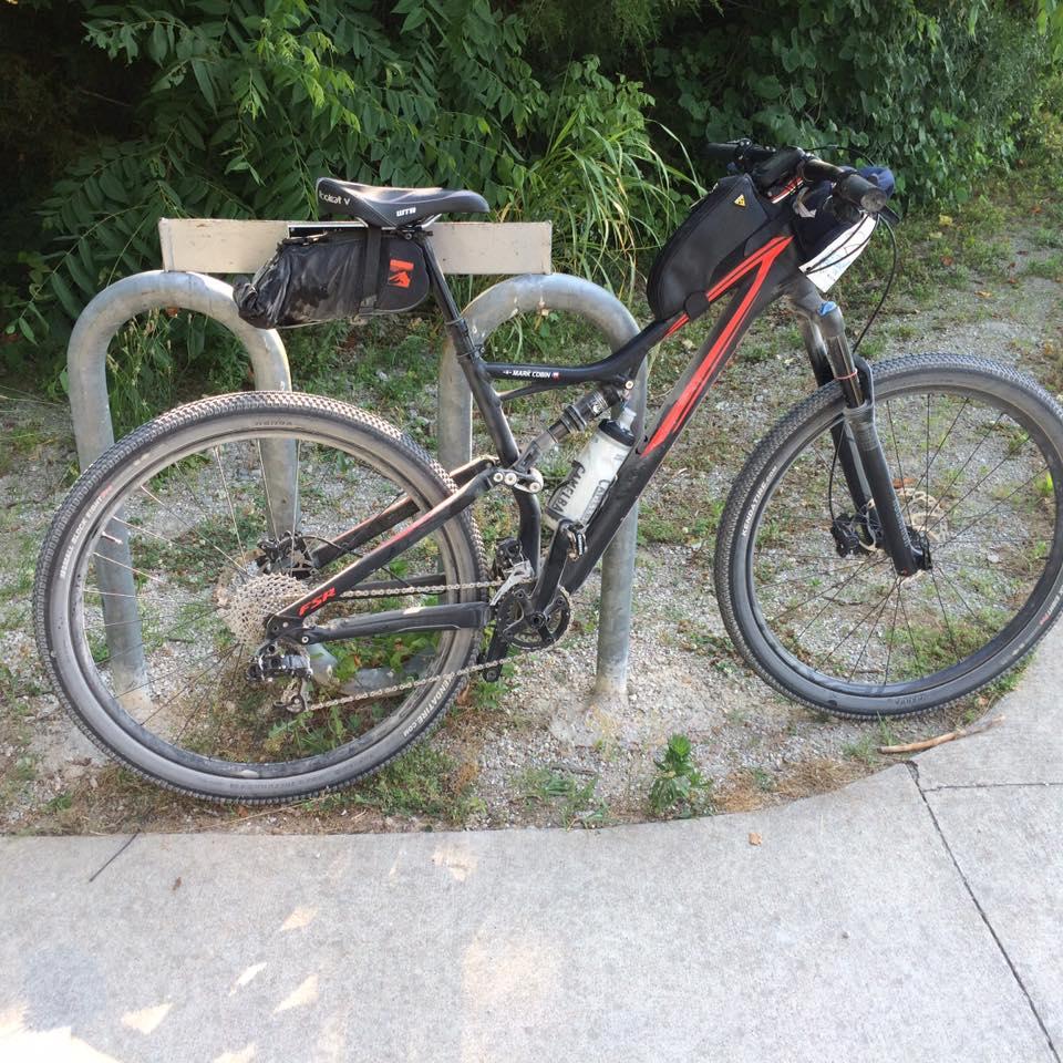Specialized Stumpjumper: A black and red mountain bike with a water bottle attached is parked at a bike rack, surrounded by green foliage. The bike features thick tires and a rear storage bag, resting on a gravel path that curves into a concrete sidewalk.