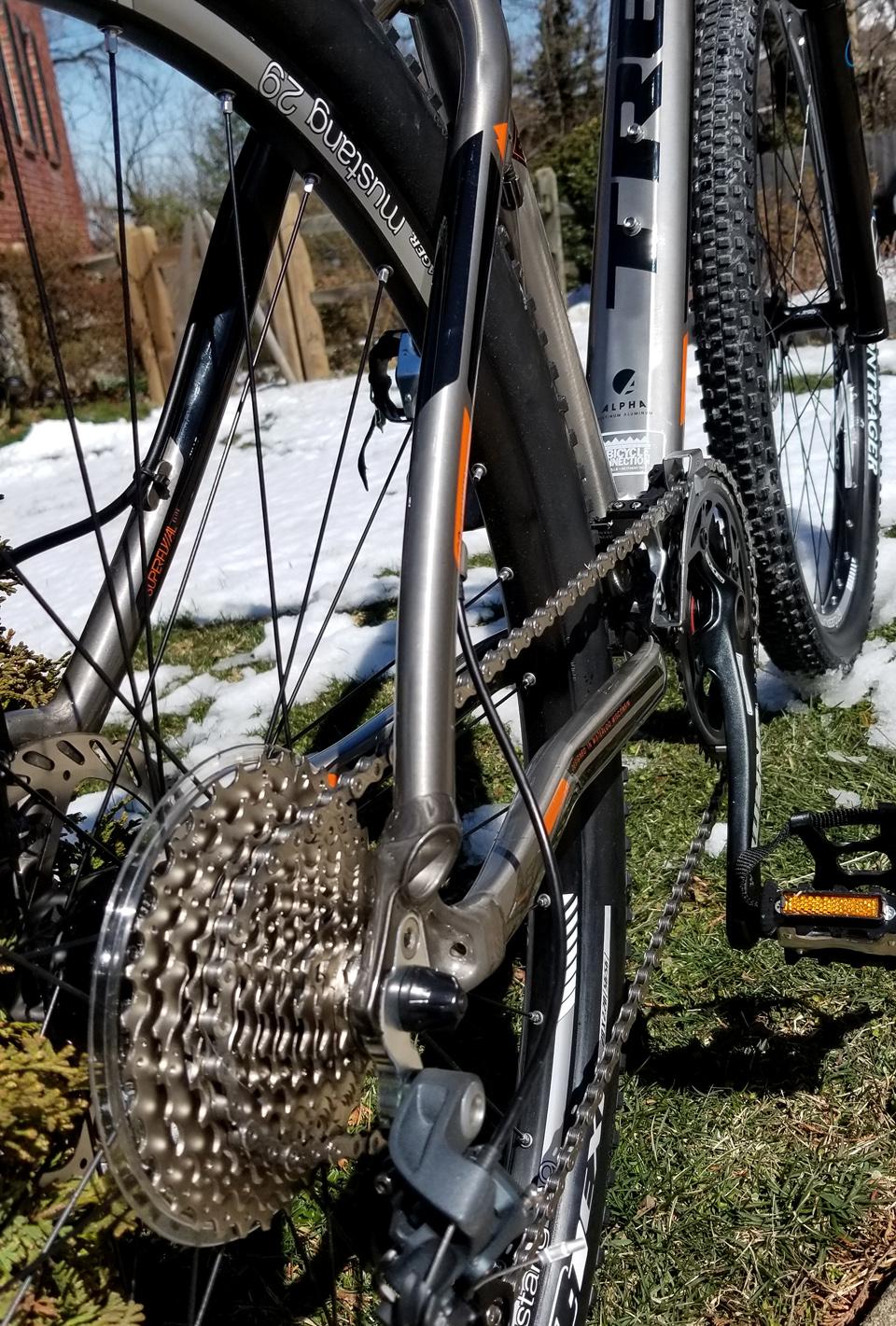 Trek Superfly AL Elite: Close-up view of the rear drivetrain and cassette of a mountain bike, showcasing the gear system and chain. The bike is positioned on grass with some patches of snow in the background, indicating early spring conditions. The frame is silver with orange accents, highlighting its modern design.