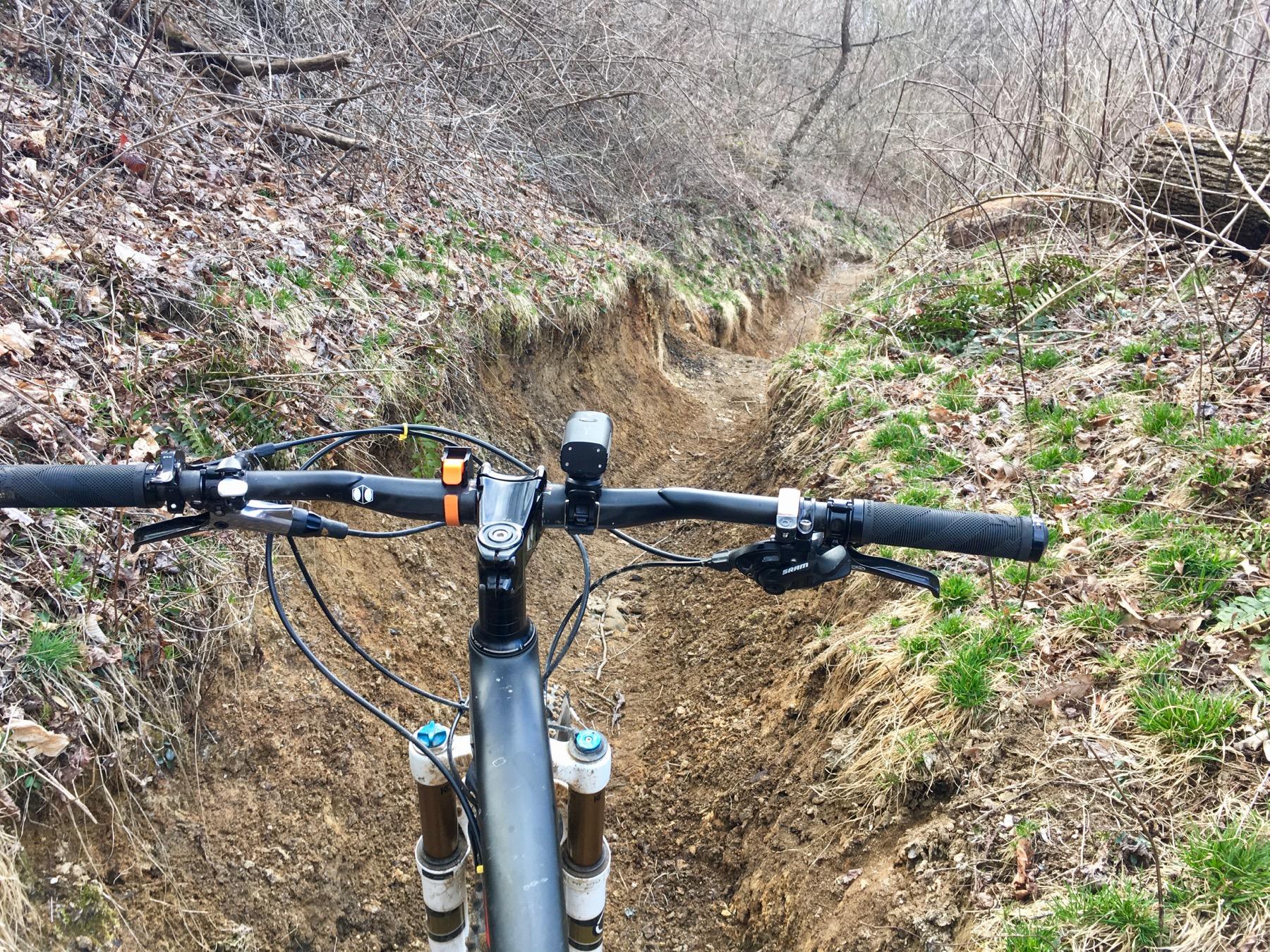 A view from the handlebars of a mountain bike, showing a narrow dirt trail surrounded by sparse vegetation and leaf litter. The trail appears to be slightly eroded, with a steep bank on one side and patches of green grass peeking through the ground. Avery Creek / 327 mountain bike trail.