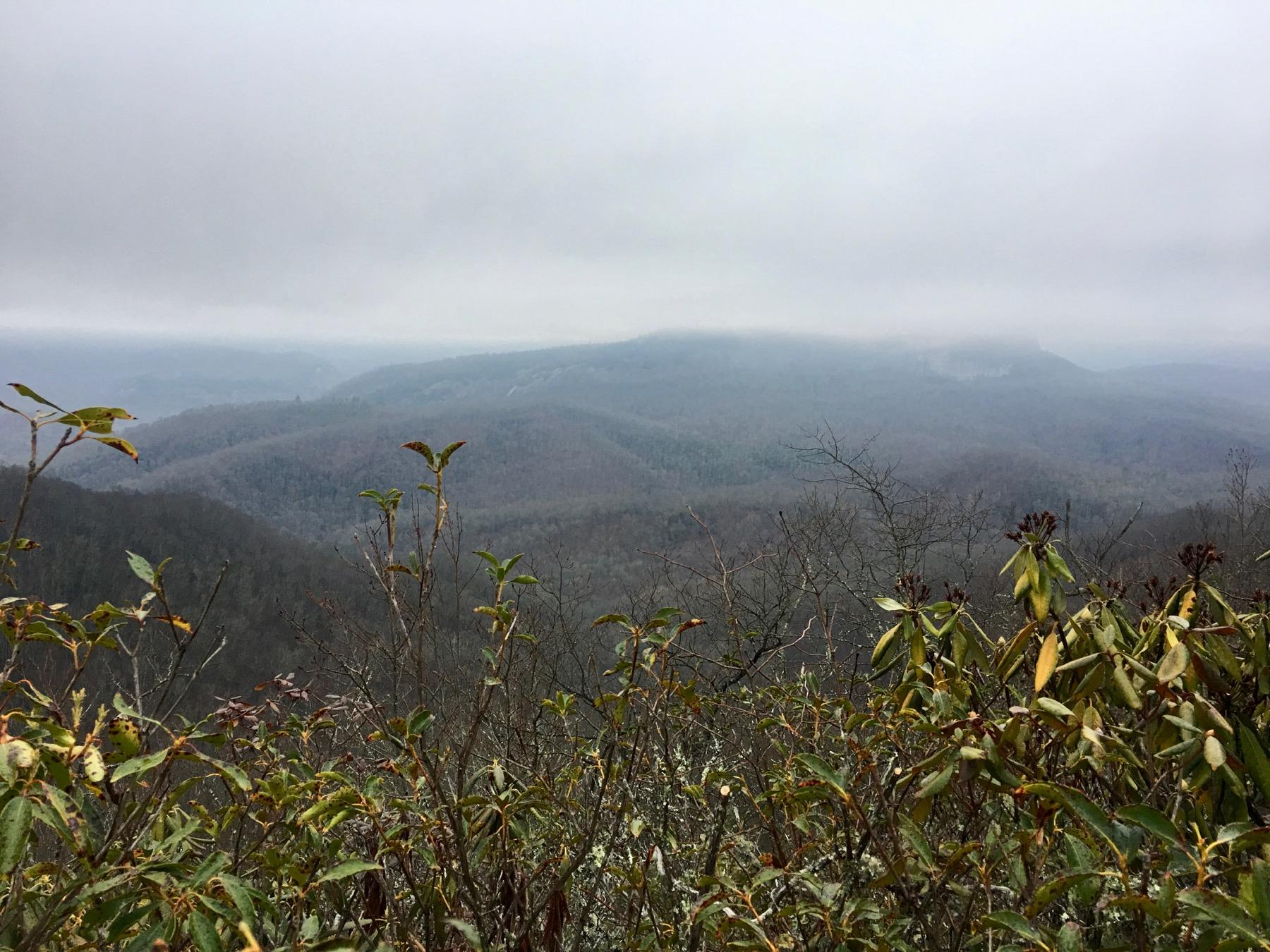 A foggy mountain landscape with layers of rolling hills covered in sparse vegetation. The foreground features green and brown foliage, while the background reveals distant mountains shrouded in mist under a cloudy sky. The scene evokes a tranquil and mysterious atmosphere. Bennett Gap / 138 mountain bike trail.