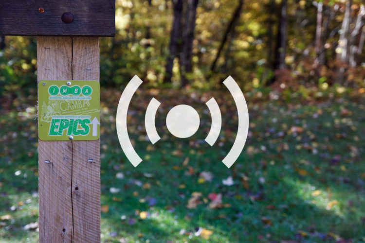 A wooden post with a green trail marker sign indicating the trail's designation as an "Epic" route by the International Mountain Bicycling Association (IMBA) and the Chequamegon Area Mountain Bike Association (CAMBA), set against a blurred background of colorful autumn foliage.