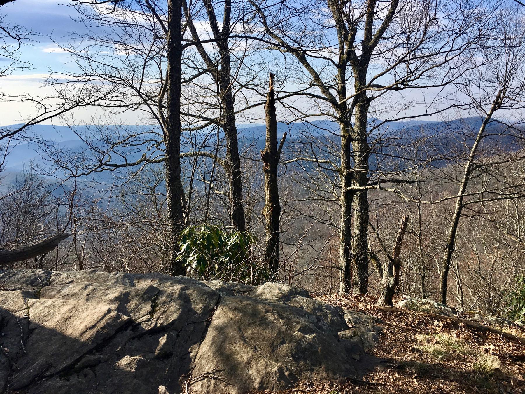 A rocky outcrop in a natural setting, surrounded by bare trees and distant mountains under a clear blue sky.  Black Mountain mountain bike trail.