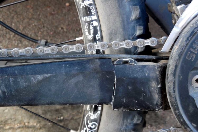 Close-up view of a bicycle's rear chain and crank area, showcasing a partially obscured bike frame, visible chain links, and a tire in the background. The image highlights details of the bike's mechanics and design.