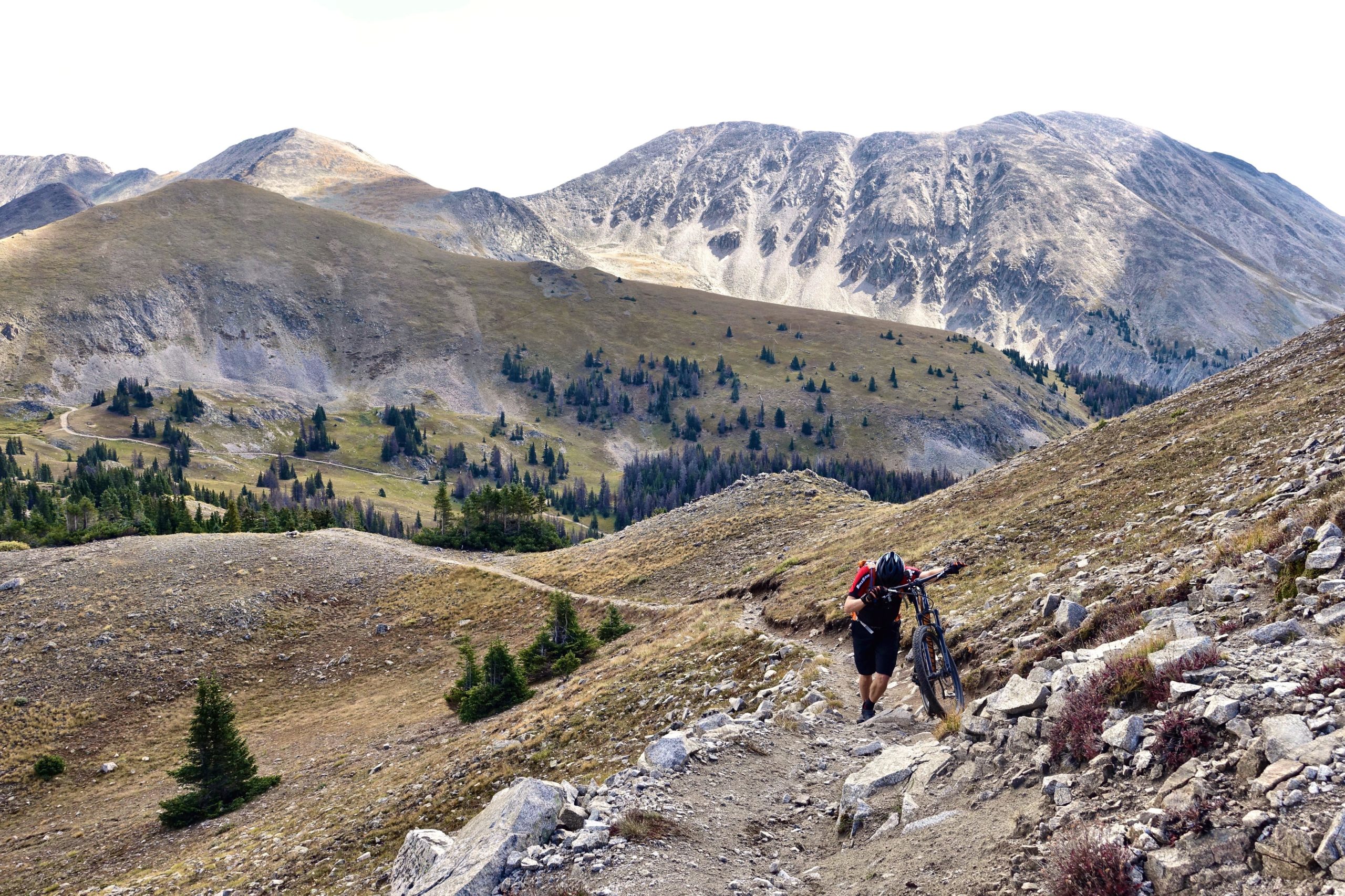 A person walking up a rocky mountain trail while carrying a mountain bike, surrounded by rolling hills and sparse vegetation, with tall mountains in the background. Canyon Creek Trail mountain bike trail.