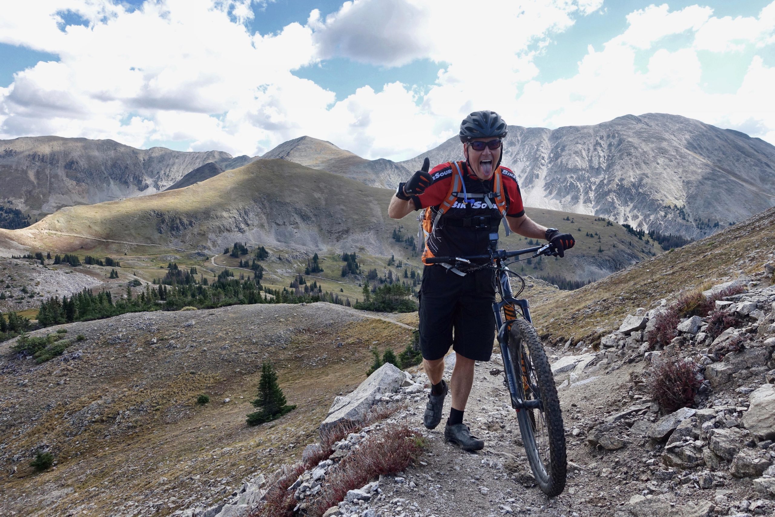 A mountain biker standing on a rocky trail in a mountainous landscape, smiling and giving a thumbs-up. The background features rolling hills and rocky peaks under a partly cloudy sky. Canyon Creek Trail mountain bike trail.