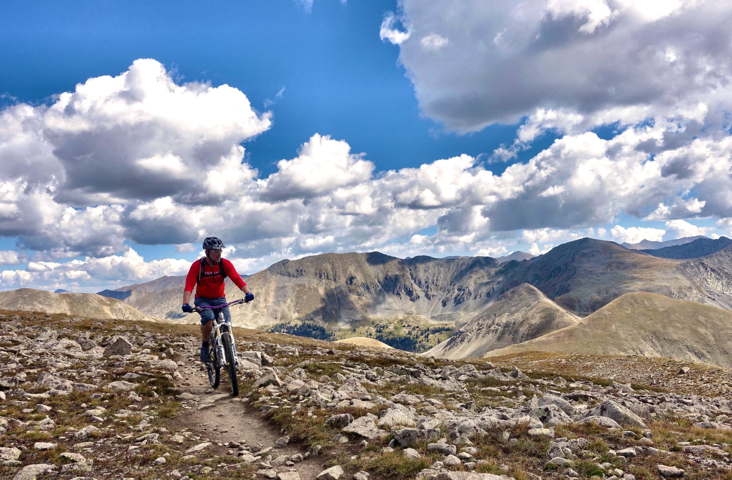 A mountain biker riding along a rocky trail in a mountainous landscape, with expansive blue skies and fluffy clouds overhead. The surrounding terrain features rolling hills and distant peaks, showcasing a scenic and adventurous outdoor environment. Canyon Creek Trail mountain bike trail.