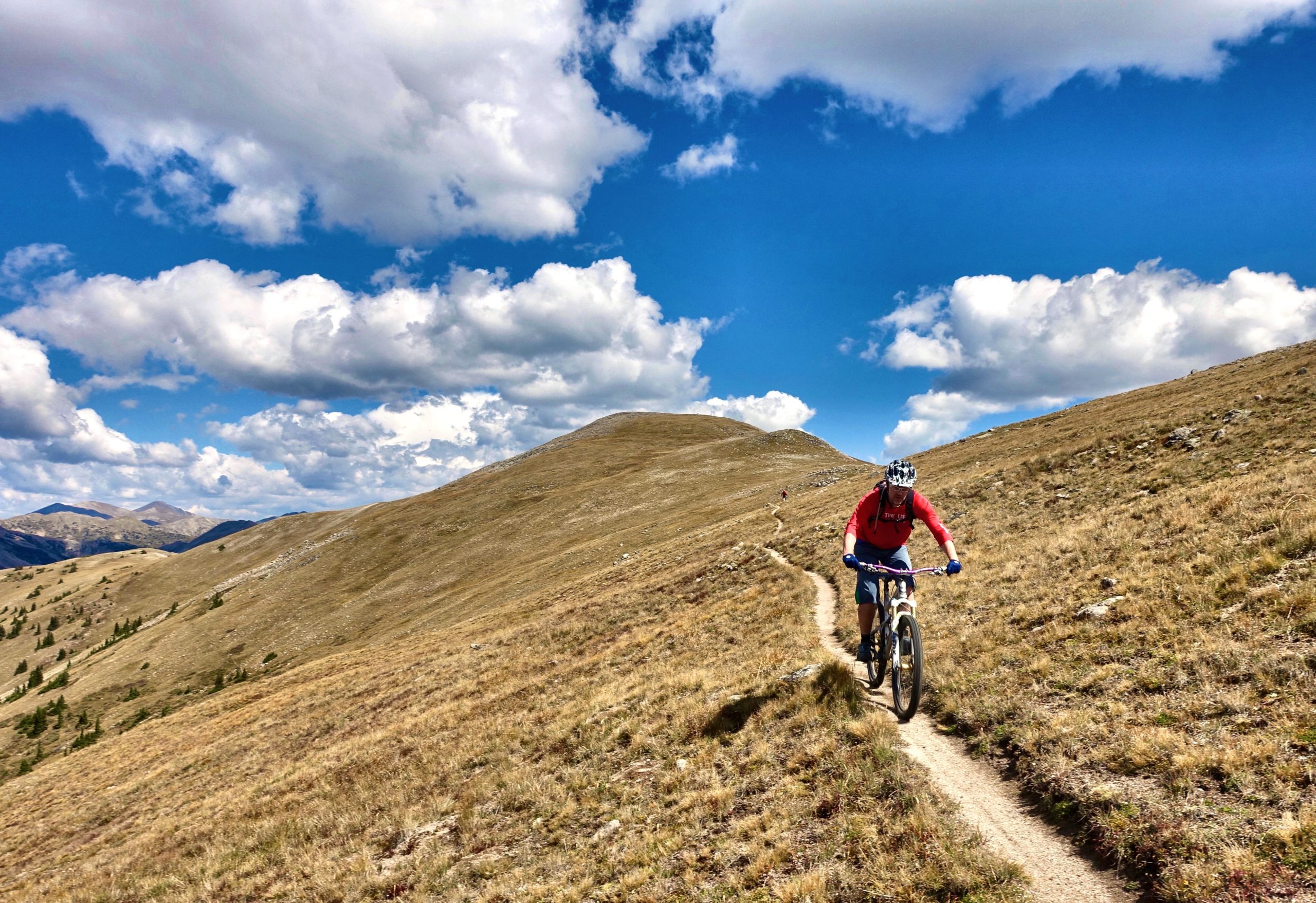 A mountain biker riding along a narrow dirt trail on a grassy hillside, with vibrant blue skies and fluffy white clouds in the background. Rolling mountains are visible in the distance, creating a scenic outdoor backdrop. Canyon Creek Trail mountain bike trail.