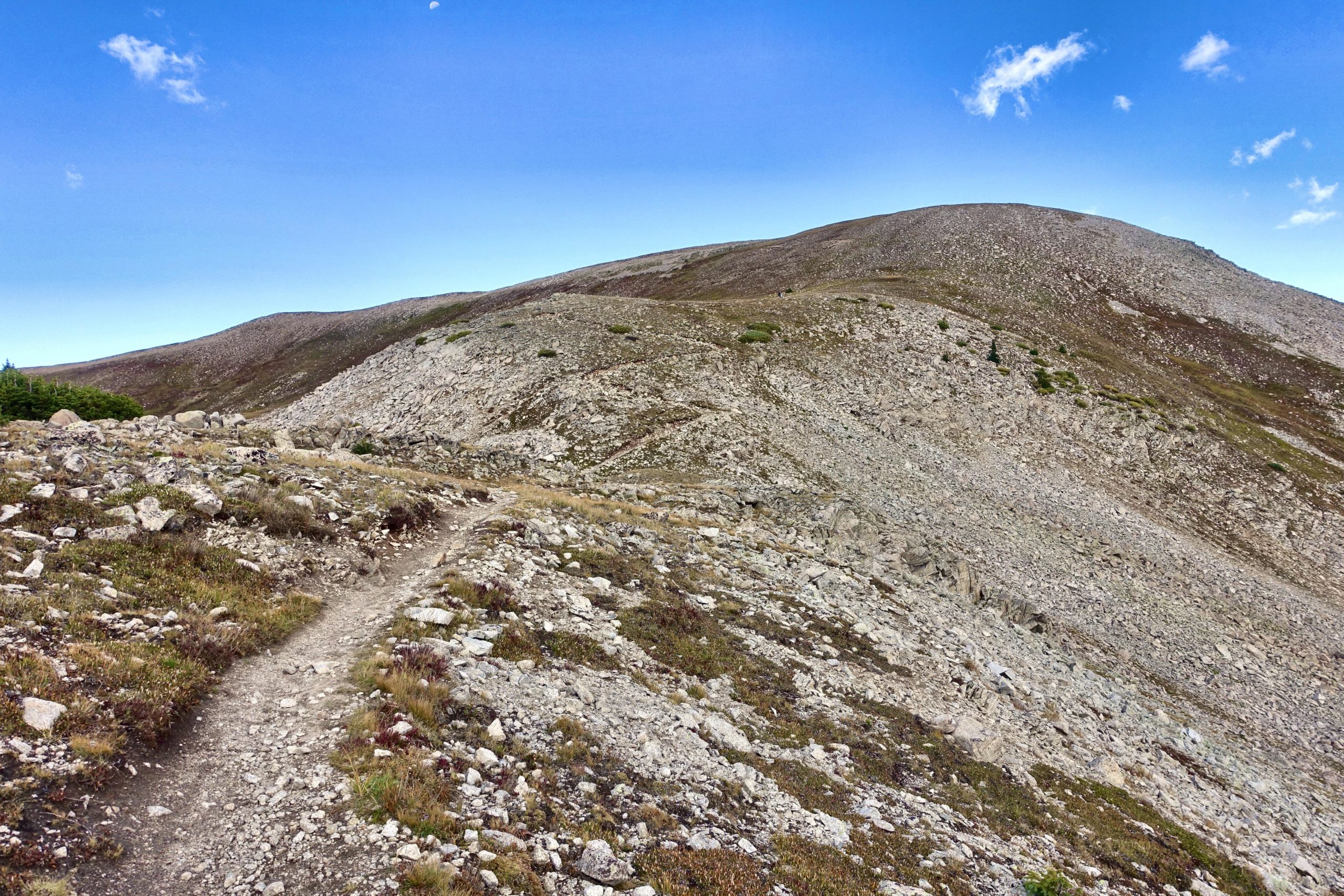 A rocky hiking trail leads through a mountainous landscape, flanked by gentle slopes and patches of greenery under a blue sky with a few scattered clouds. The terrain is rugged, with boulders and sparse vegetation visible along the path, suggesting a natural, wilderness setting. Canyon Creek Trail mountain bike trail.