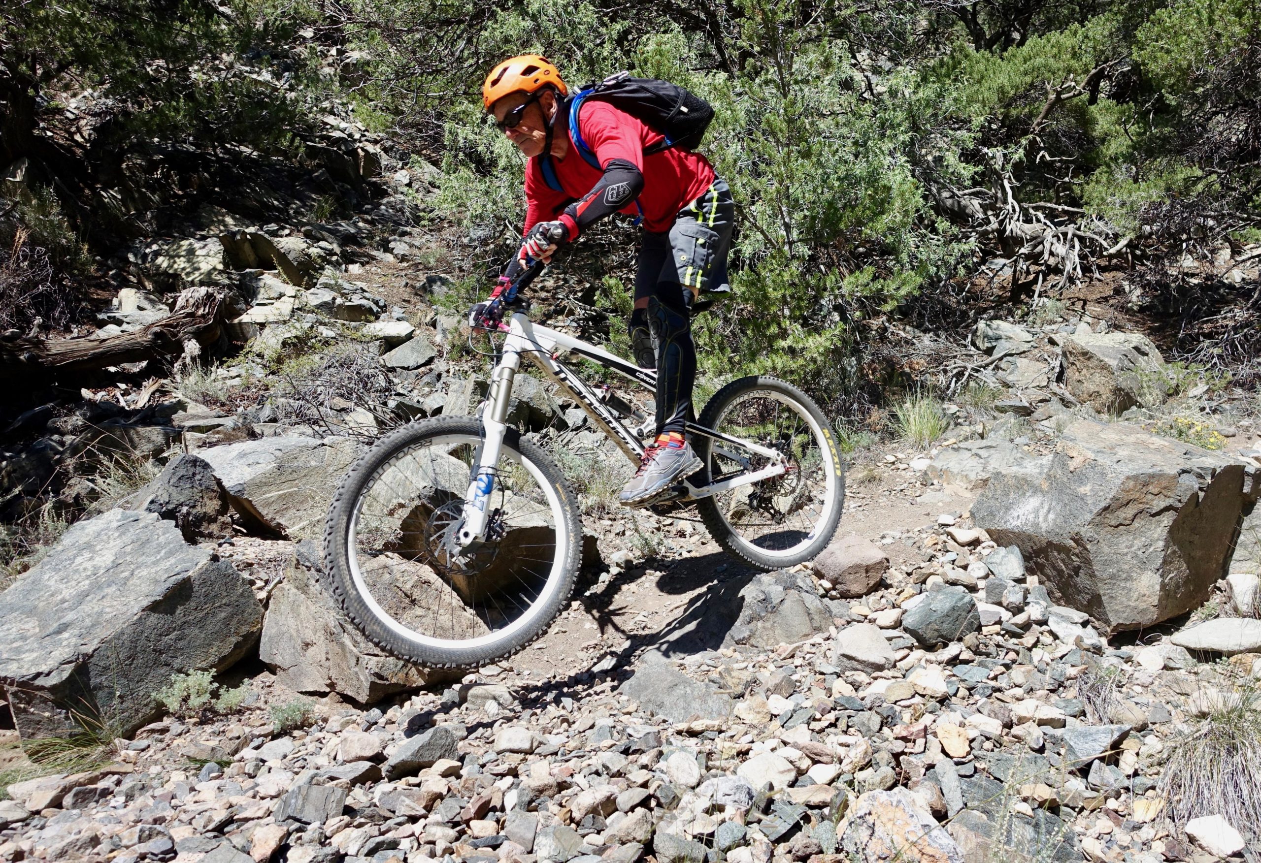 A mountain biker in protective gear, including an orange helmet and sunglasses, jumps over rocks on a challenging trail surrounded by trees and dense foliage. The rider is captured mid-air, showcasing the excitement of off-road biking. Cottonwood mountain bike trail.