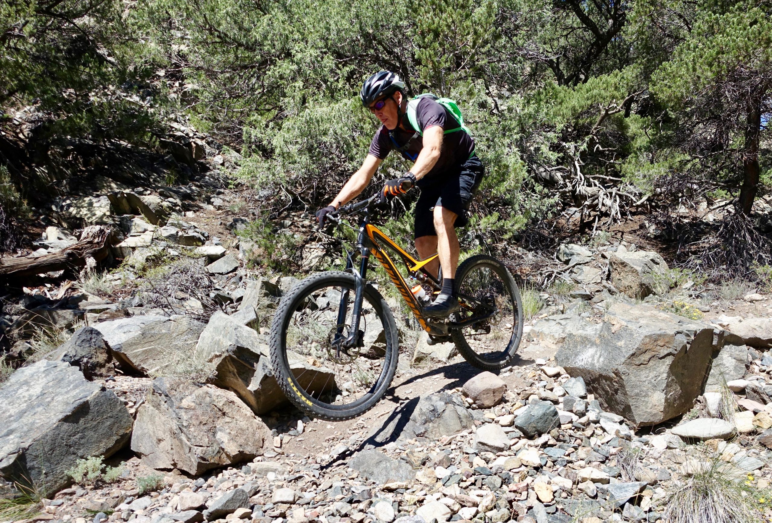 A mountain biker riding over rocky terrain in a wooded area. The rider is focused as they navigate the challenging path, surrounded by greenery and boulders. Cottonwood mountain bike trail.