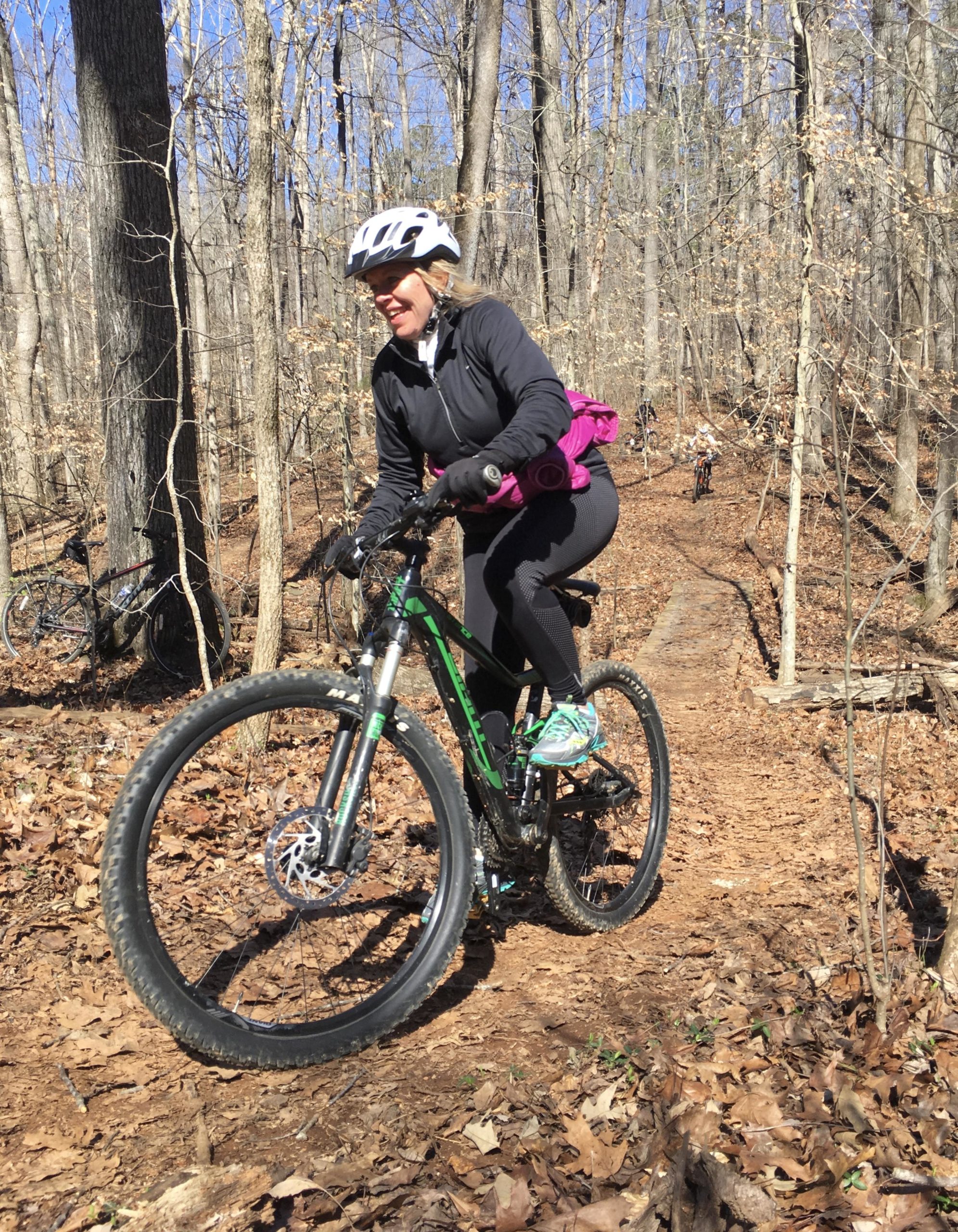 A woman rides a mountain bike on a dirt trail surrounded by bare trees. She is wearing a helmet and athletic clothing, with a backpack slung over her shoulder. The trail is covered in fallen leaves, and another cyclist can be seen in the background. The scene captures a sunny day in a forested area, emphasizing the outdoor activity and nature. Tanglewood Park mountain bike trail.