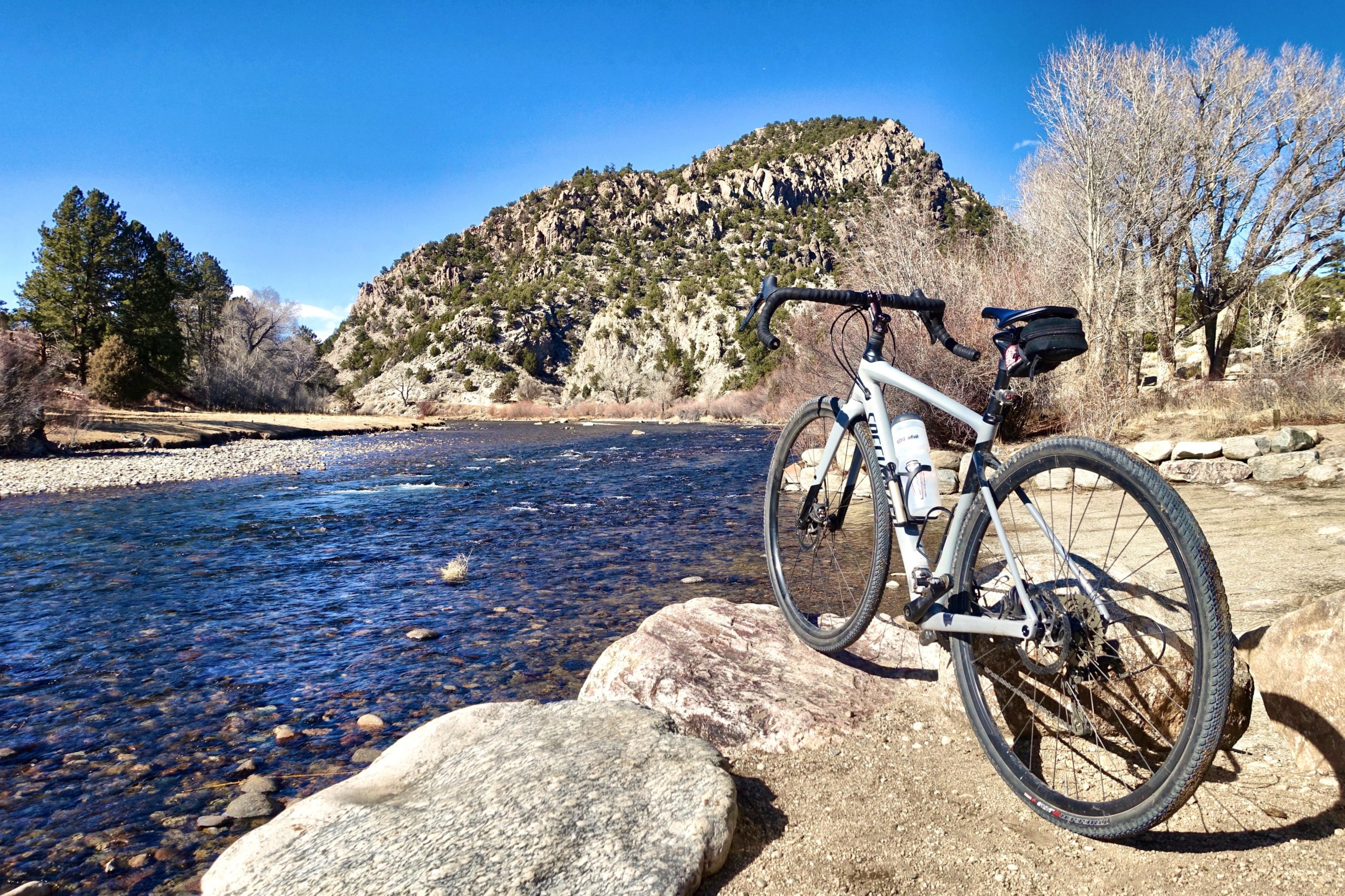 A gray bicycle is parked on a rocky shore by a clear, flowing river, with mountains and pine trees in the background under a bright blue sky. Bald Mountain Road / #300 mountain bike trail.