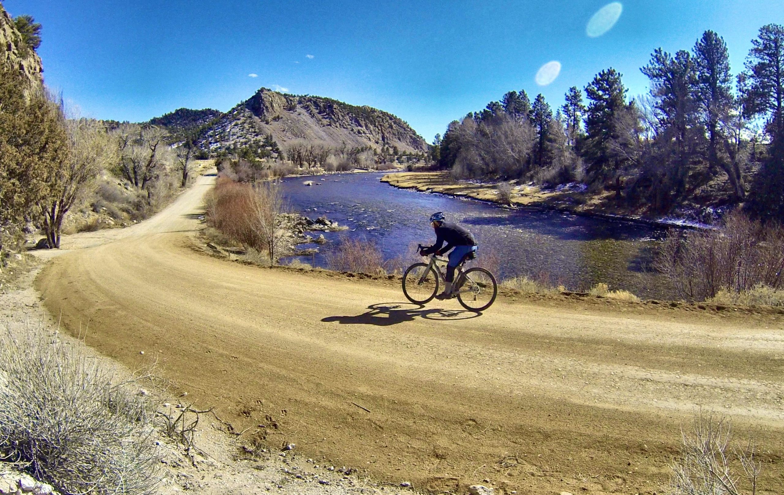 A cyclist riding on a dirt trail alongside a river, surrounded by trees and mountains under a clear blue sky. The path curves gently, and the sunlight casts shadows on the ground. Bald Mountain Road / #300 mountain bike trail.