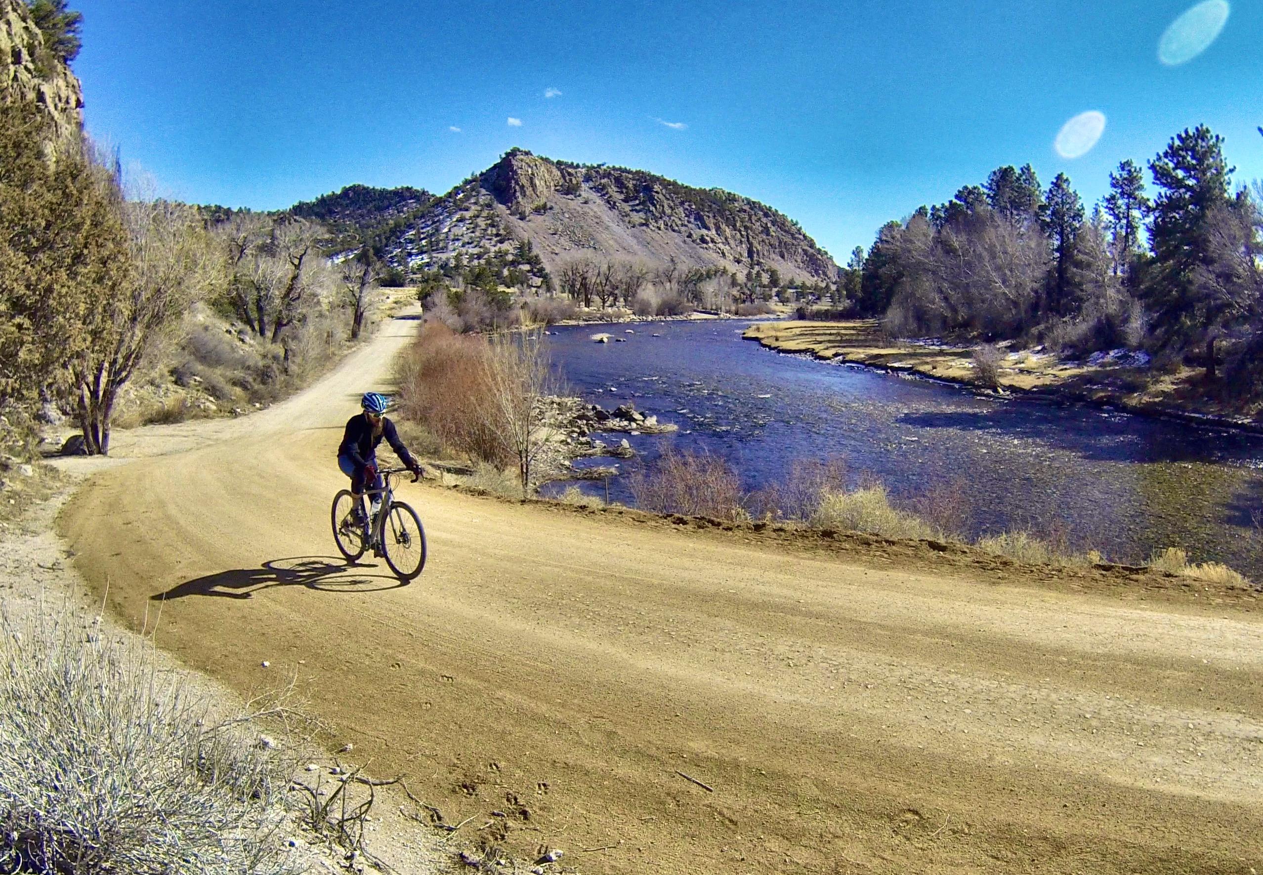 A cyclist riding a bike along a dirt path next to a river, surrounded by trees and rocky hills under a clear blue sky. Bald Mountain Road / #300 mountain bike trail.