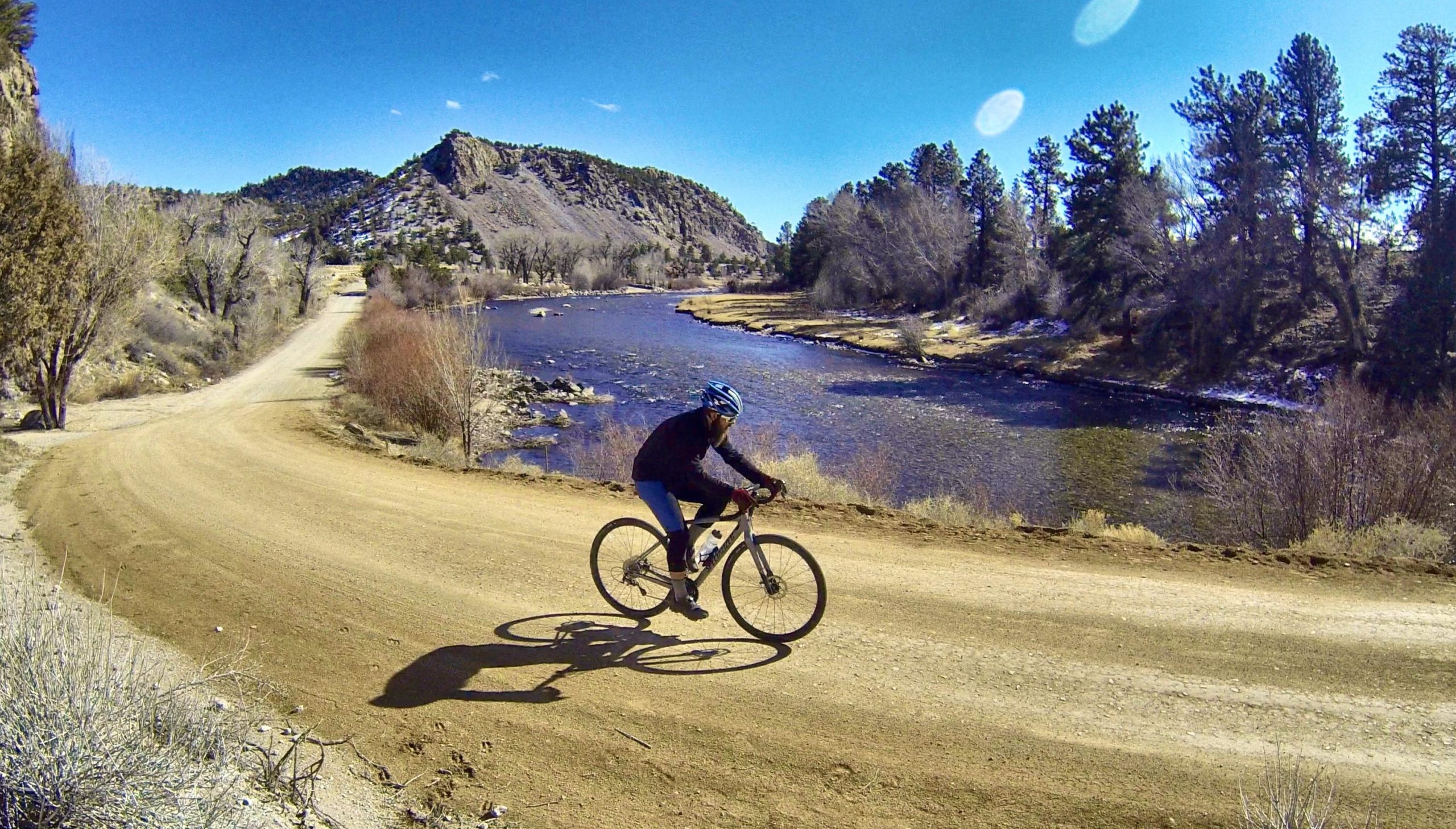 A cyclist rides on a dirt path along a river, surrounded by trees and hills under a clear blue sky. The scene depicts a peaceful outdoor environment, showcasing the beauty of nature and recreational biking. Bald Mountain Road / #300 mountain bike trail.