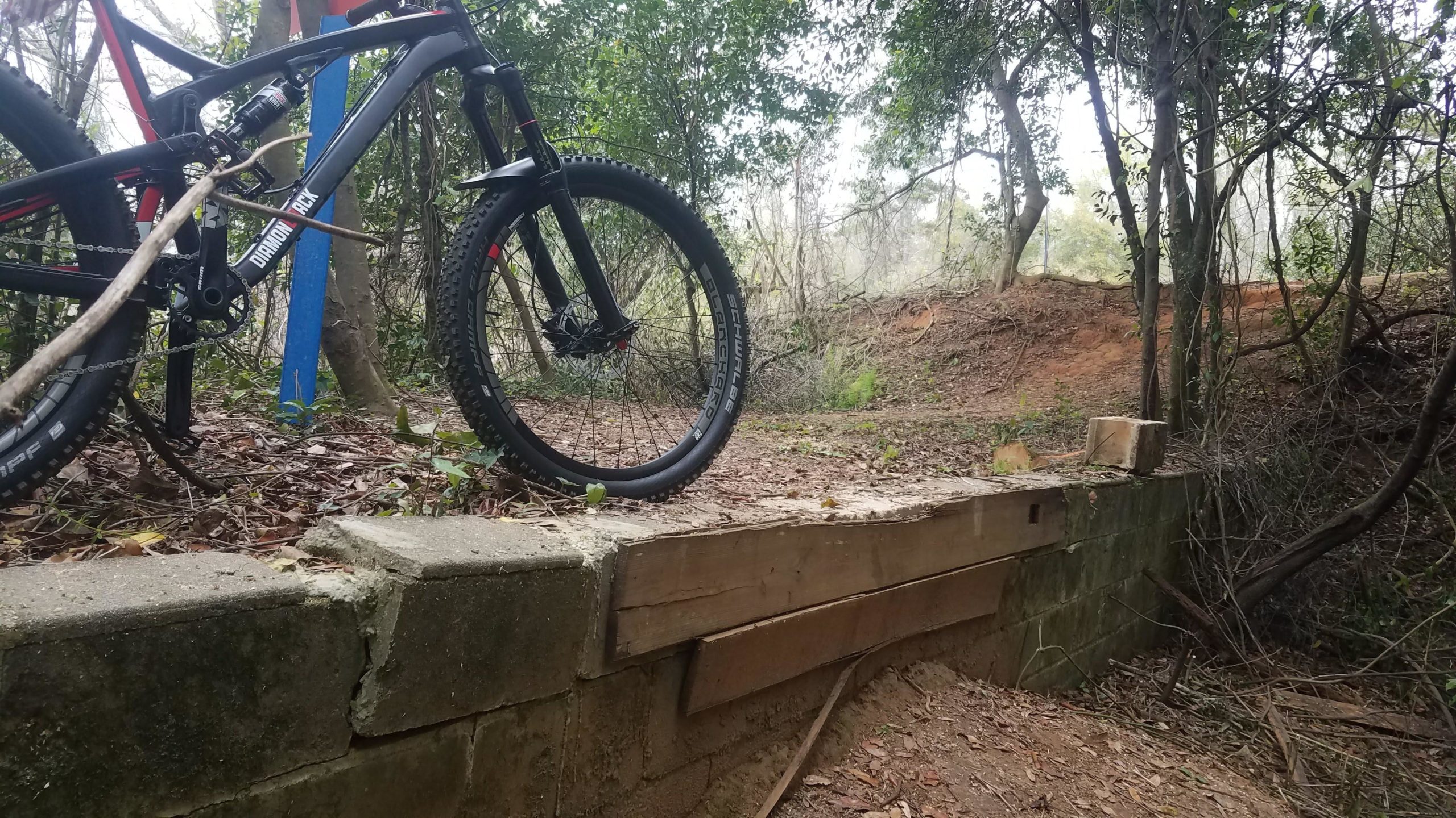 A mountain bike leaning against a concrete and wooden structure in a wooded area, with dense foliage in the background and a trail visible in the distance. Tom Brown / Lafayette Heritage Park mountain bike trail.
