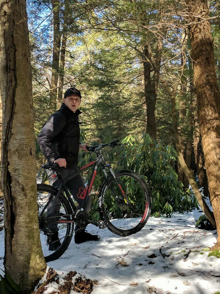 Jamis Durango 29er: A person standing on snow-covered ground in a forest, holding a mountain bike. The surrounding trees are lush and green, indicating a natural setting, with patches of snow visible on the ground. The person is wearing a black jacket and a hat, looking slightly towards the camera.