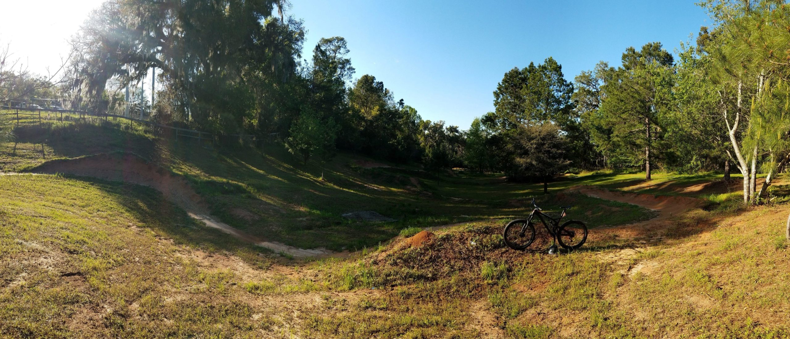 A panoramic view of a grassy, tree-lined trail designed for biking, with a black mountain bike resting on a small dirt mound in the foreground. The scene is illuminated by sunlight filtering through the trees, highlighted by clear blue skies and shadows on the ground. The landscape features gentle slopes and a natural, rustic appearance, suitable for outdoor recreation. Tom Brown / Lafayette Heritage Park mountain bike trail.