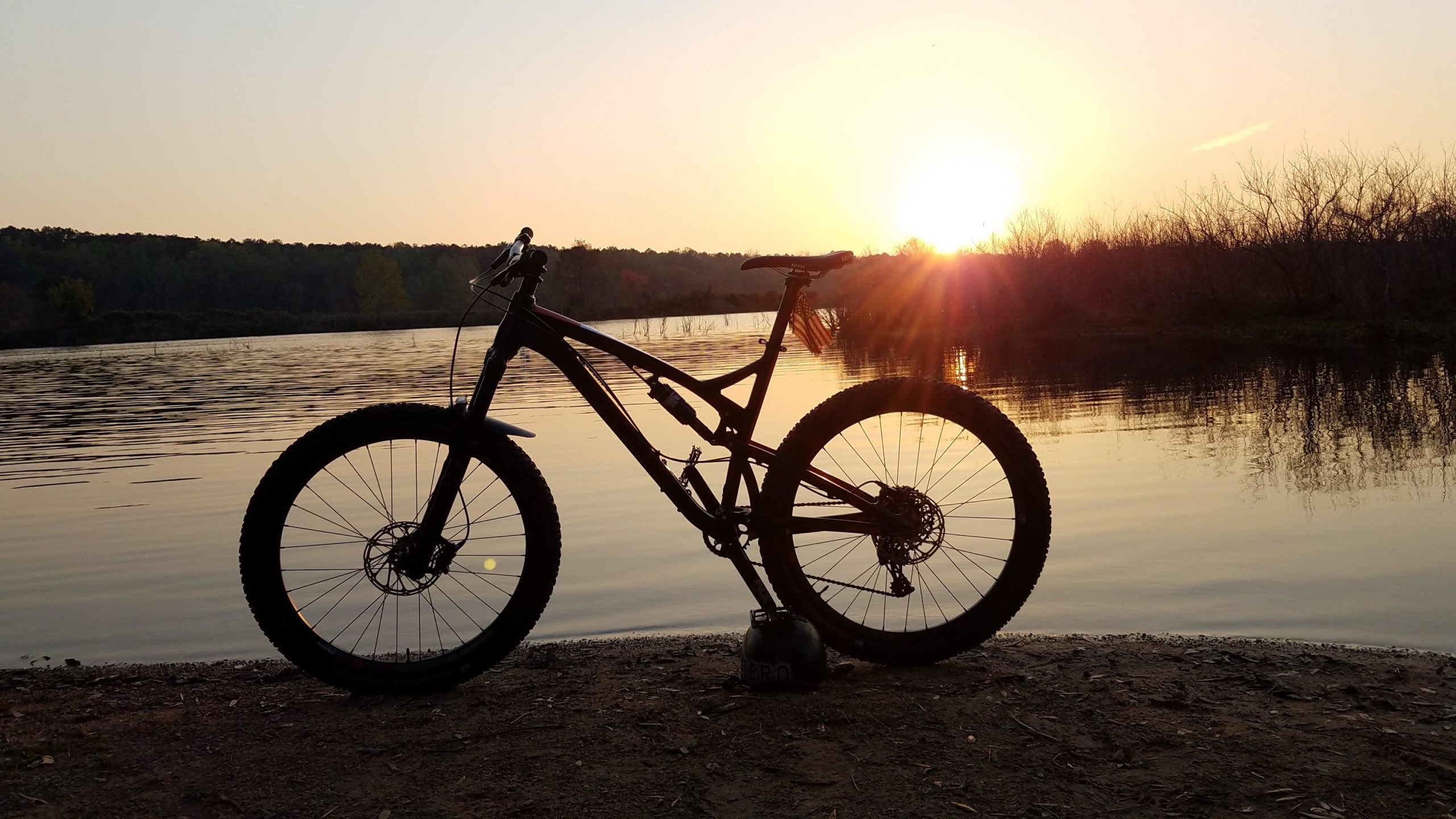 A mountain bike silhouetted against a sunset over a calm lake, with reflections on the water and trees in the background. A bike helmet rests on the sandy shore. Tom Brown / Lafayette Heritage Park mountain bike trail.