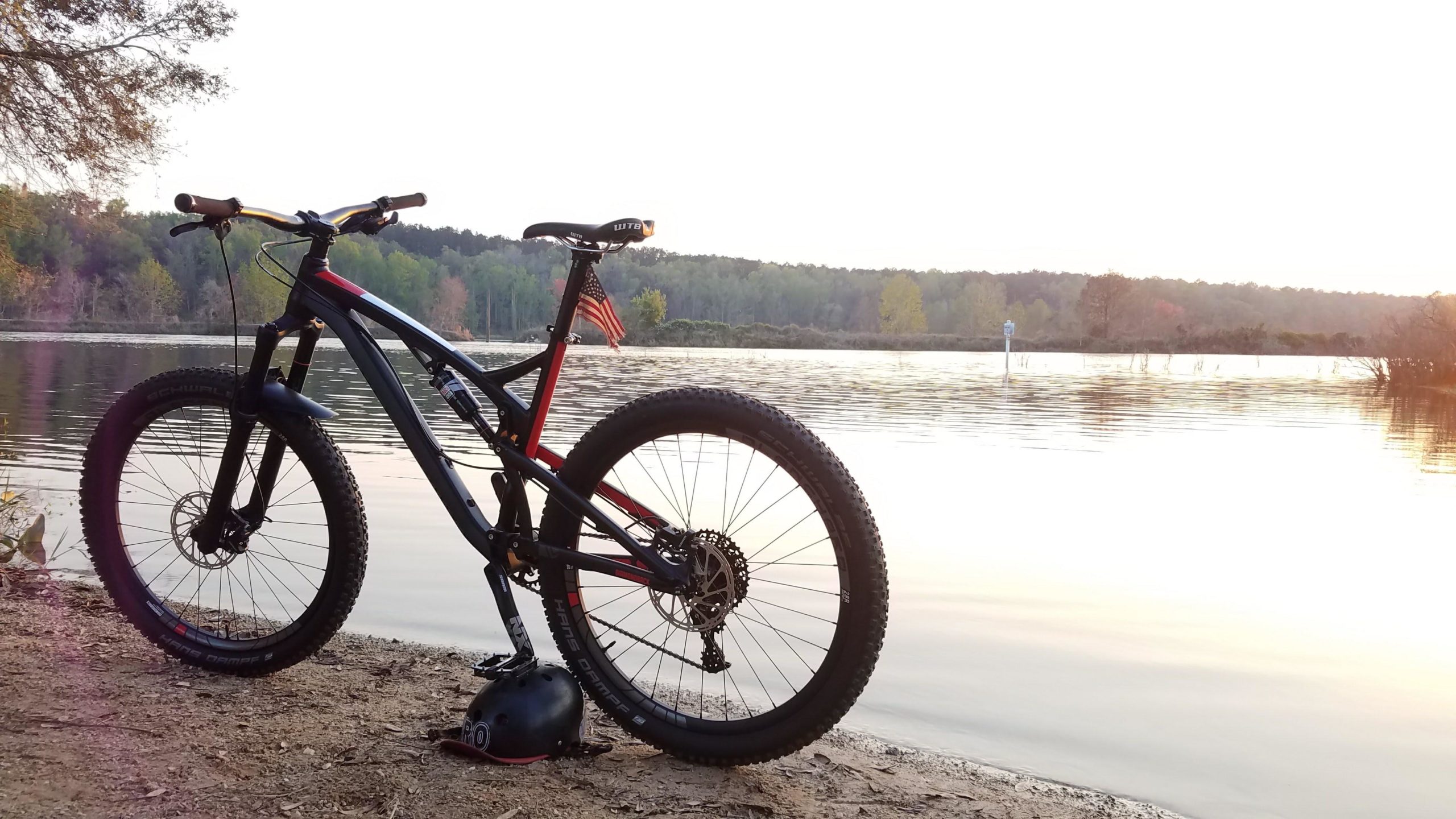 A mountain bike resting on the sandy shore of a lake at sunset, with water reflecting the fading light. A black helmet sits beside the bike, and an American flag is attached to the bike seat. Lush green trees line the opposite shore, creating a peaceful natural backdrop. Tom Brown / Lafayette Heritage Park mountain bike trail.
